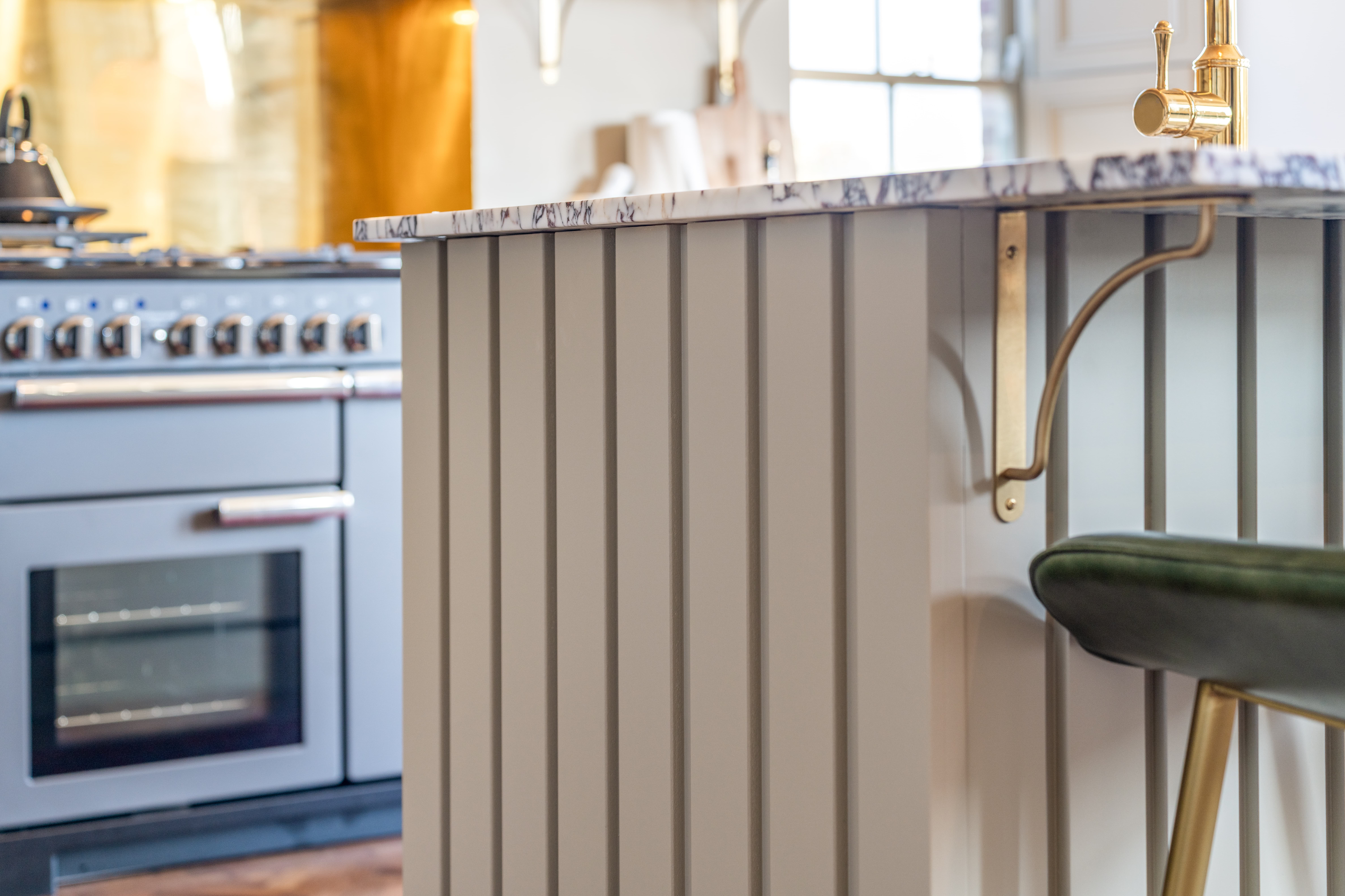 Close-up of a kitchen island with marble countertop and gold fixtures with stove in background.