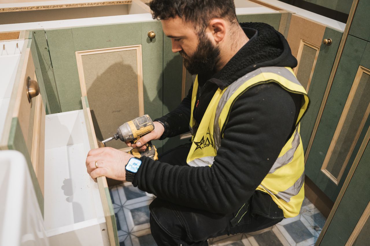 Man in yellow safety vest using power drill to assemble a green kitchen cabinet drawer.