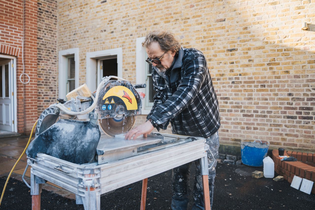 Man wearing glasses cutting tile with a wet saw outside a brick building.