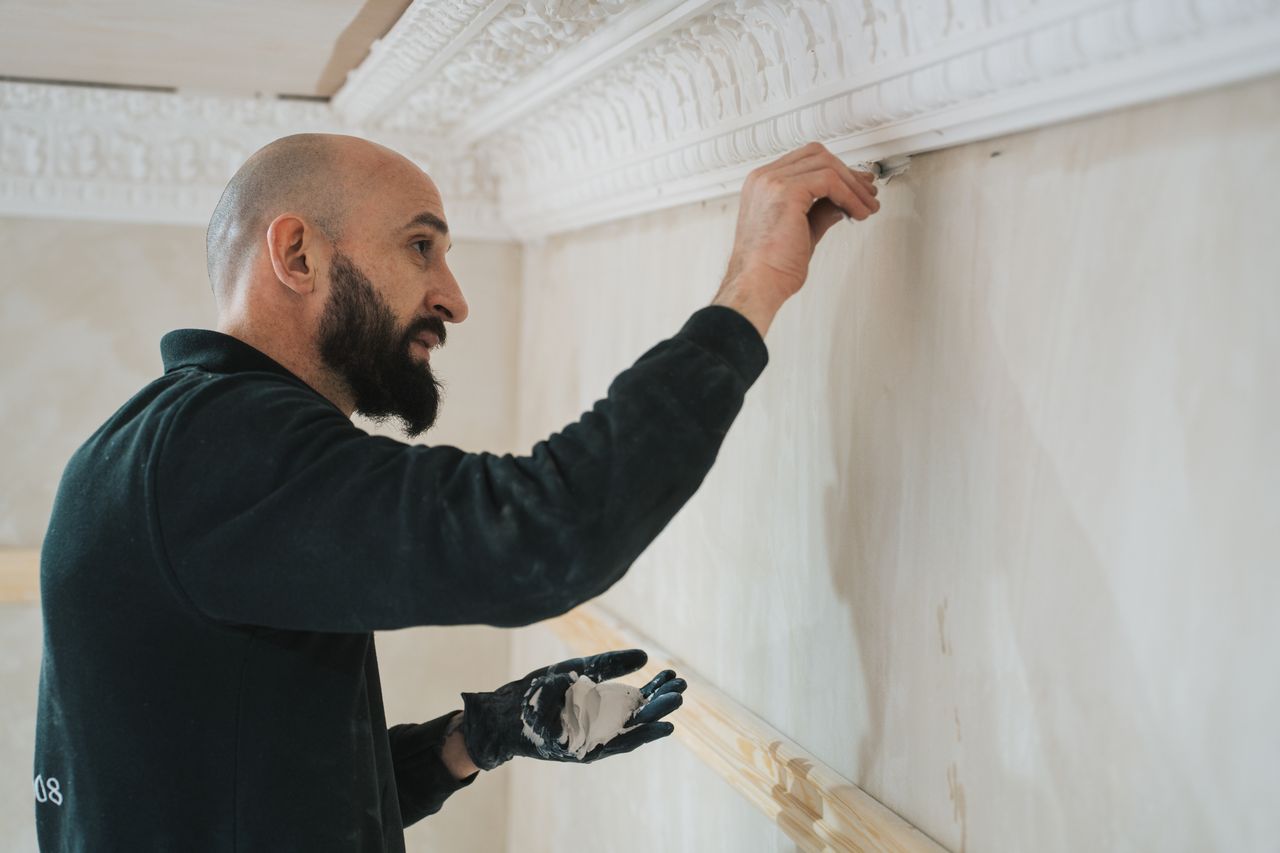 Man with beard applying plaster or paint on wall near decorative molding wearing a black glove.