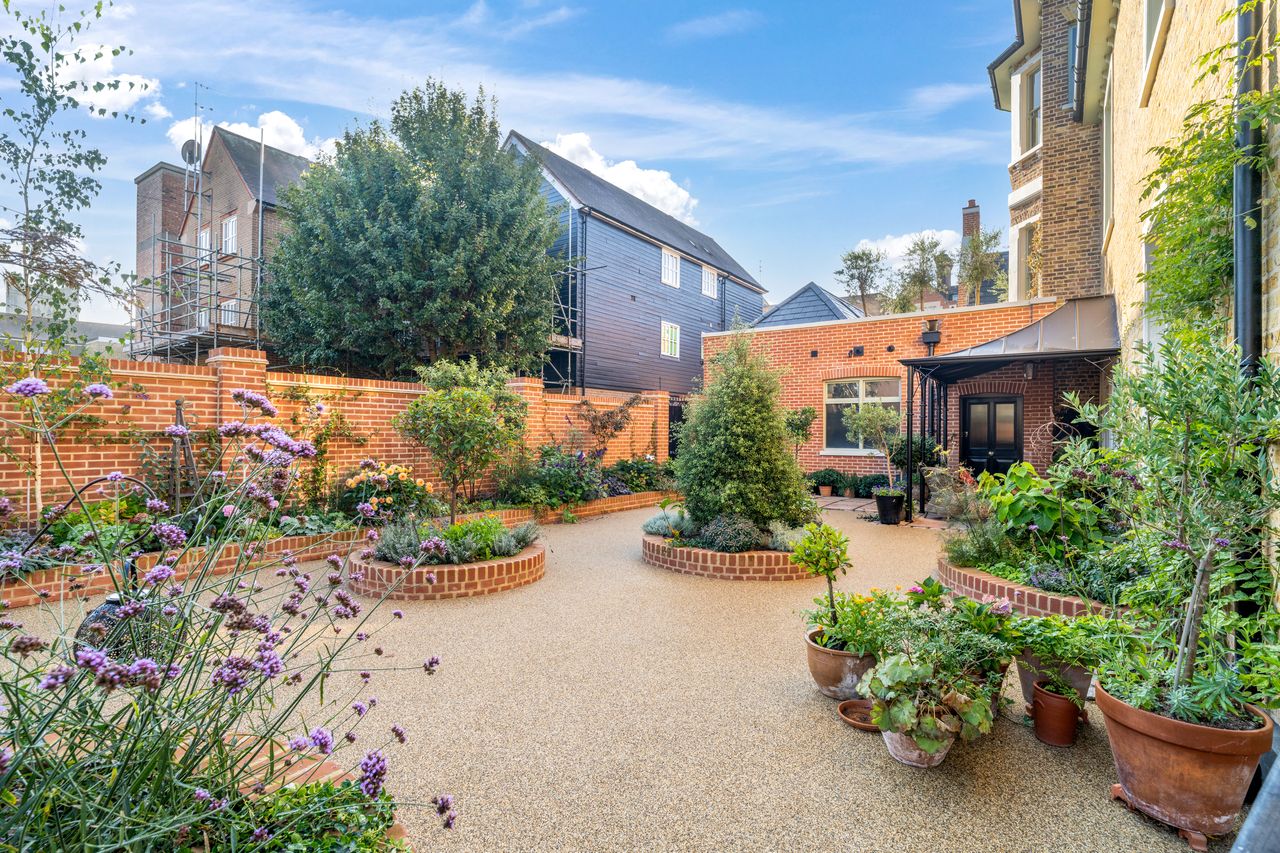 Flower garden courtyard with brick planters, potted plants, and surrounding brick buildings under blue sky.