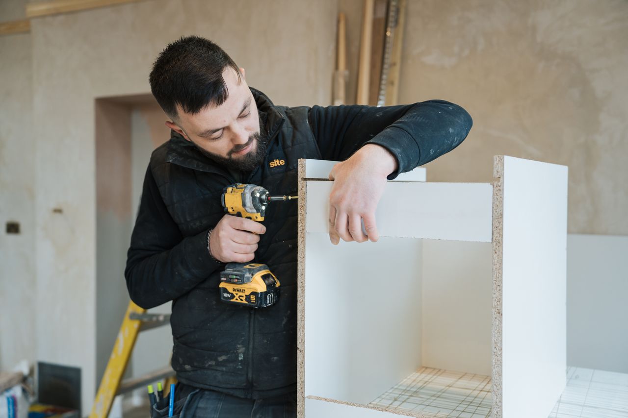 Man using a yellow power drill to assemble white wooden furniture inside a room under renovation.