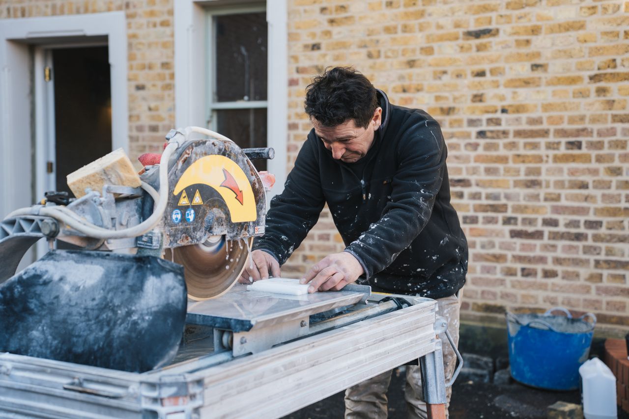 Man using a wet saw to cut a white tile outside a brick building under construction.
