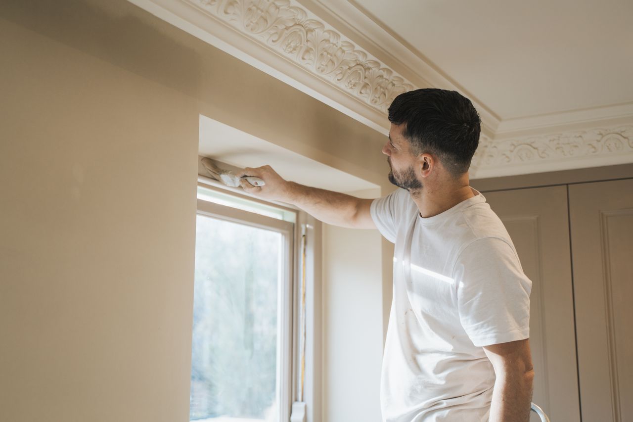 Man painting beige wall around a window inside a room with decorative white molding.