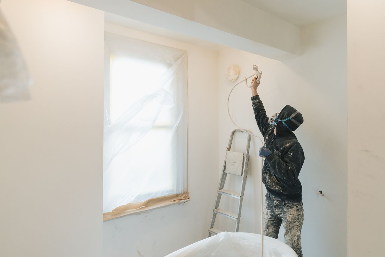 Painter wearing mask using spray gun to paint a white wall in a covered room with a ladder nearby.