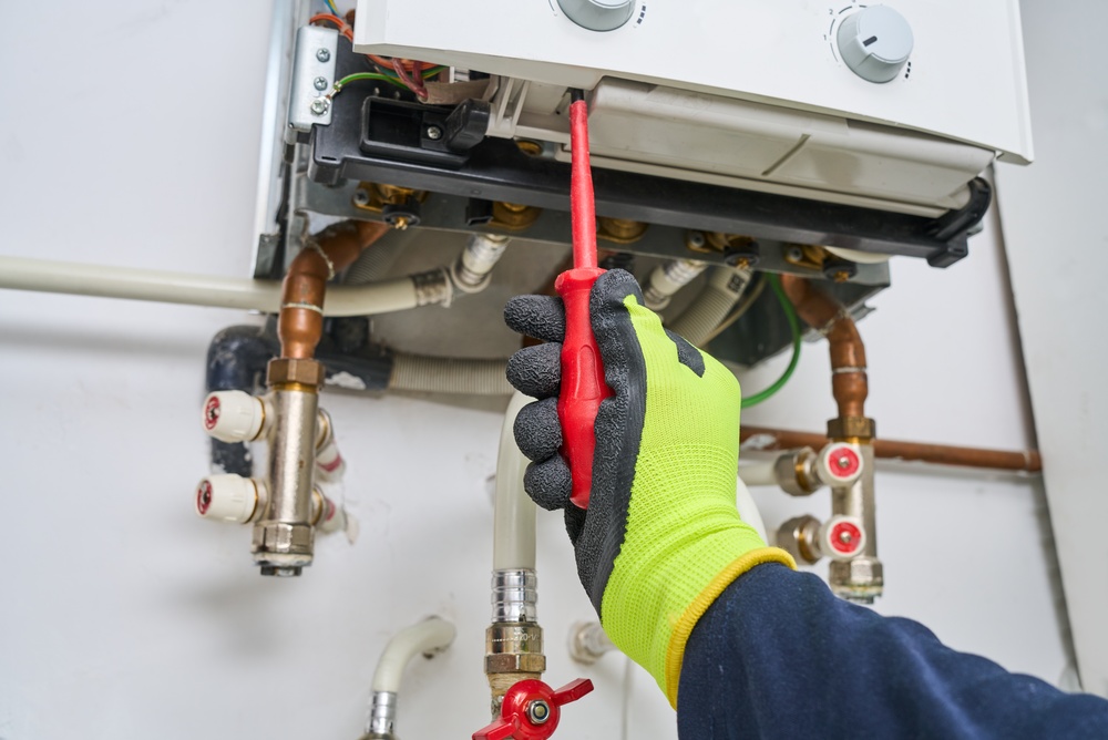 Worker in high-visibility glove using red screwdriver on heating system pipes and controls.