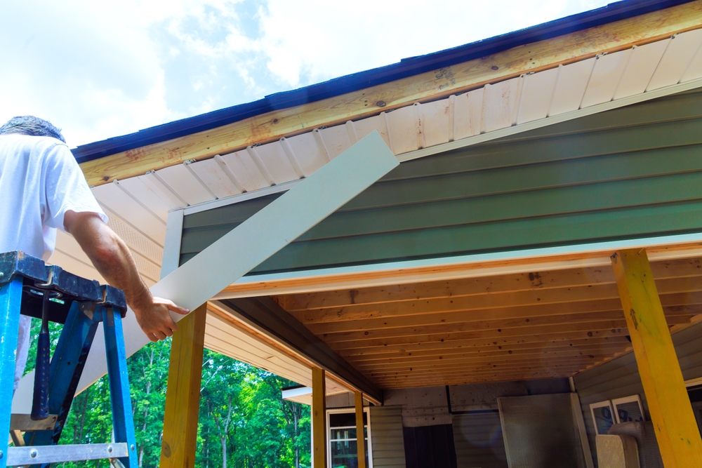 Man on ladder installing white fascia board on house roof overhang on a sunny day.