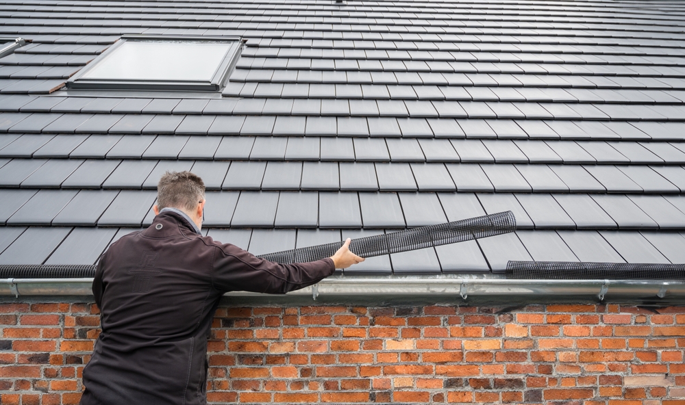 Man installing black gutter guard netting on a house gutter beneath a tiled roof with a skylight.