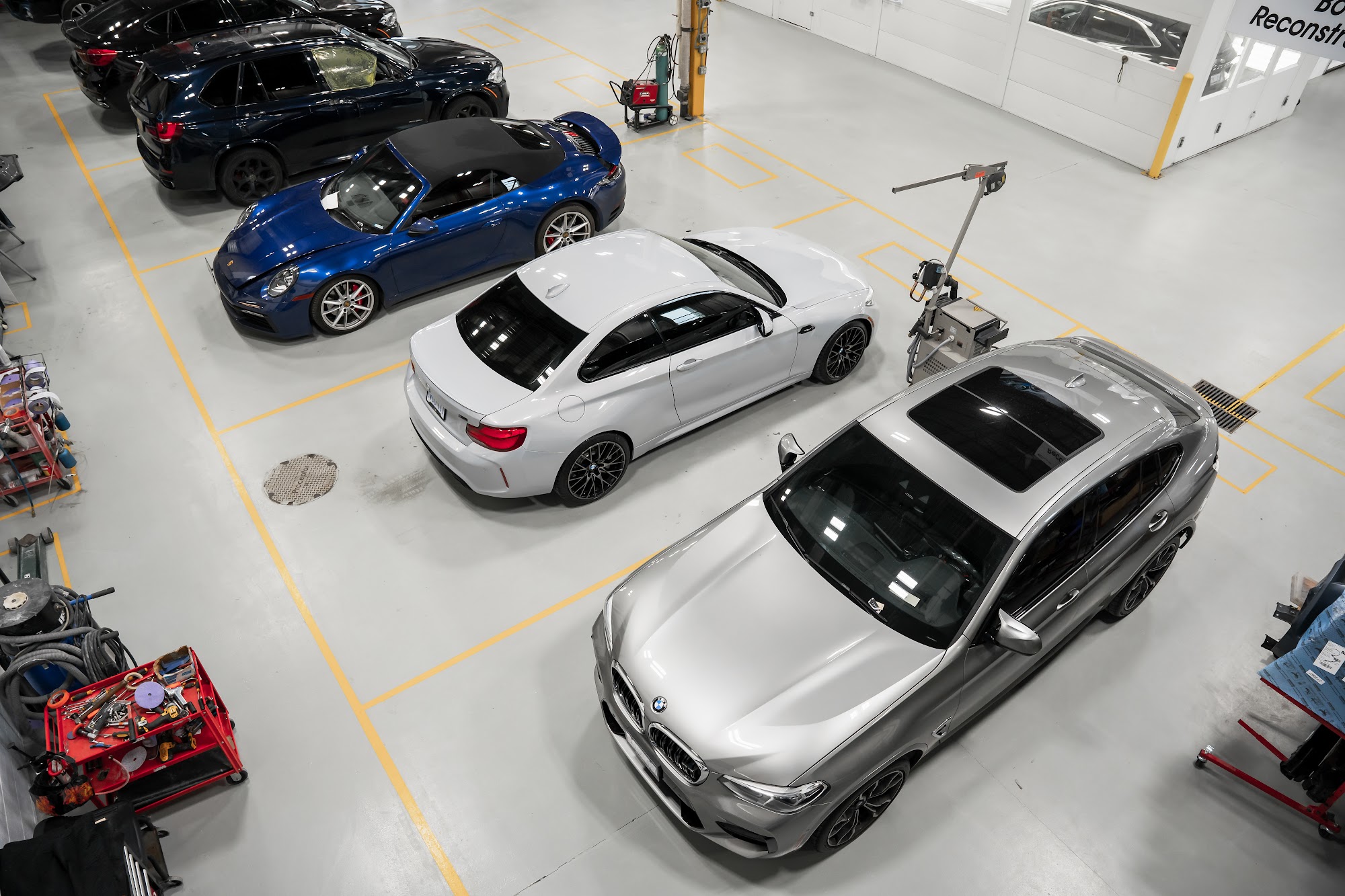 Overhead view of a blue Porsche 911, a white BMW M2 and a grey BMW X4 parked in marked bays on the floor of a certified collision centre