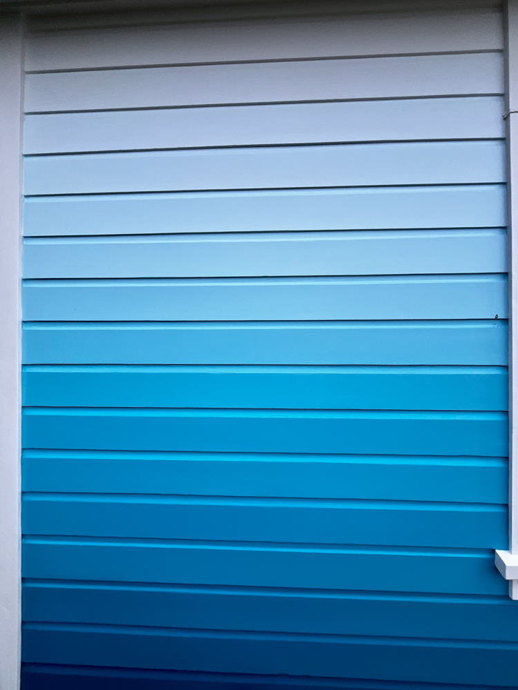 Wall siding painted in horizontal stripes showing a gradient from light blue at the top to dark blue at the bottom.
