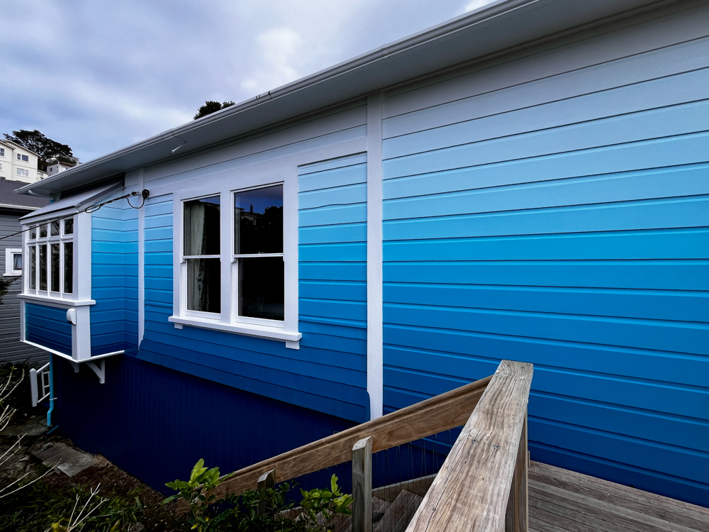 Exterior side view of a house with horizontal wooden siding painted in a gradient from dark blue at the bottom to light blue and white at the top, with two white-framed windows and a wooden handrail stairway leading down.