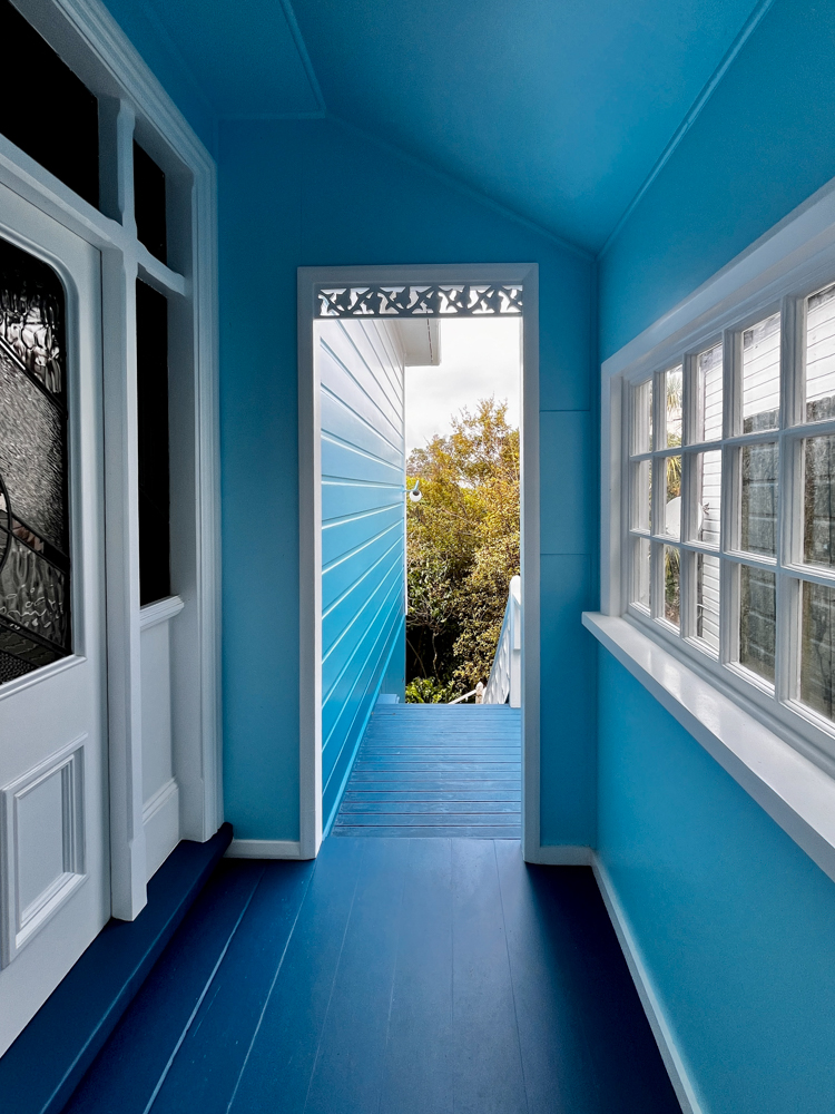 Blue-painted narrow corridor with dark blue floorboards leading to an open doorway that reveals outdoor stairs and green trees outside, with white-trimmed windows and door on sides.