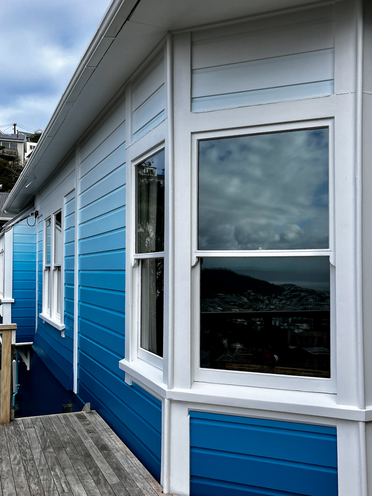Side view of a house with white framed windows and horizontal siding painted in gradient shades of blue from dark at the bottom to light at the top, with a wooden deck railing on the left.