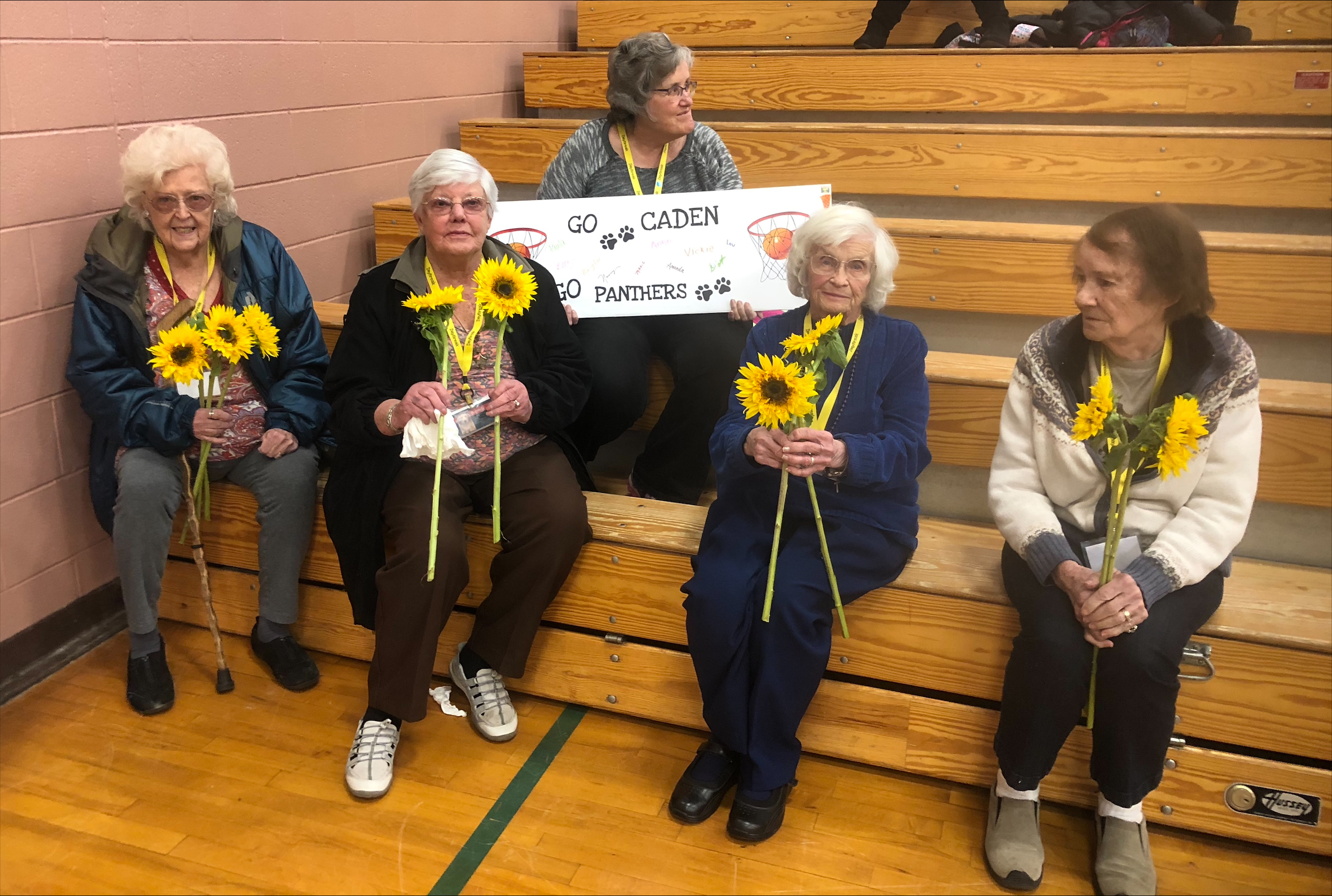Five senior women holding yellow sunflowers sitting on bleachers with a Caden Panthers banner