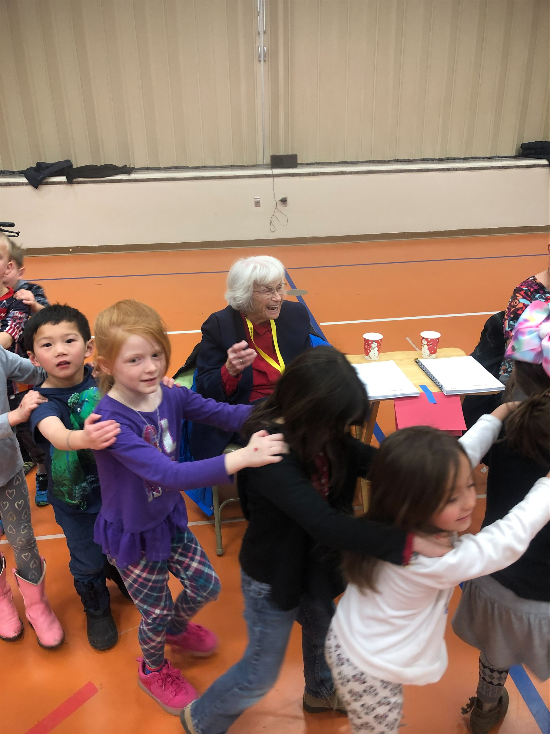Elderly woman with white hair and lanyard speaking to young children in a gymnasium