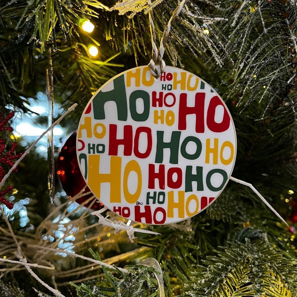 Closeup of a circular ornament hanging on a Christmas tree with the words "Ho Ho Ho" repeating multiple times to create a pattern-like effect in the design.