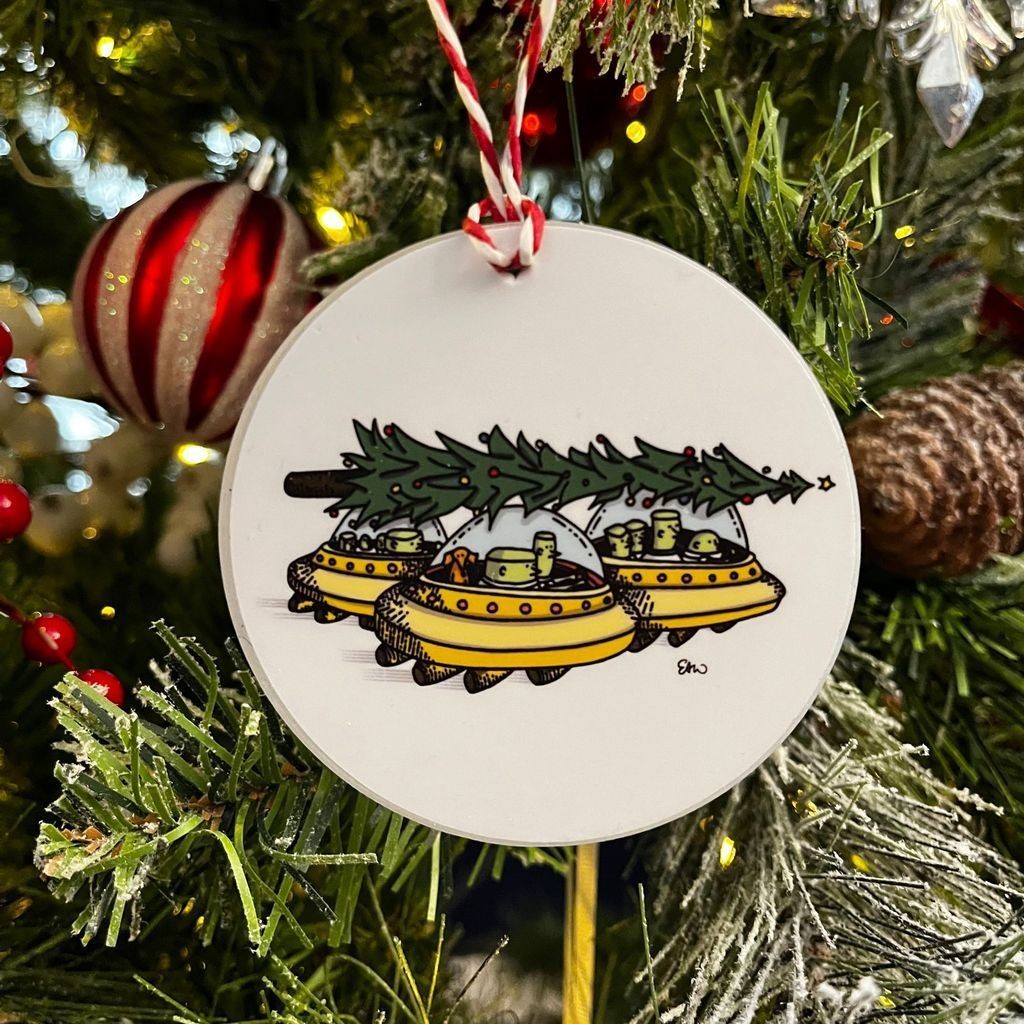 A closeup photos of an ornament hanging on a Christmas tree branch. The illustration features sitting in three flying cars that look UFOs. They have a Christmas tree strapped across the roofs of the threee flying cars.