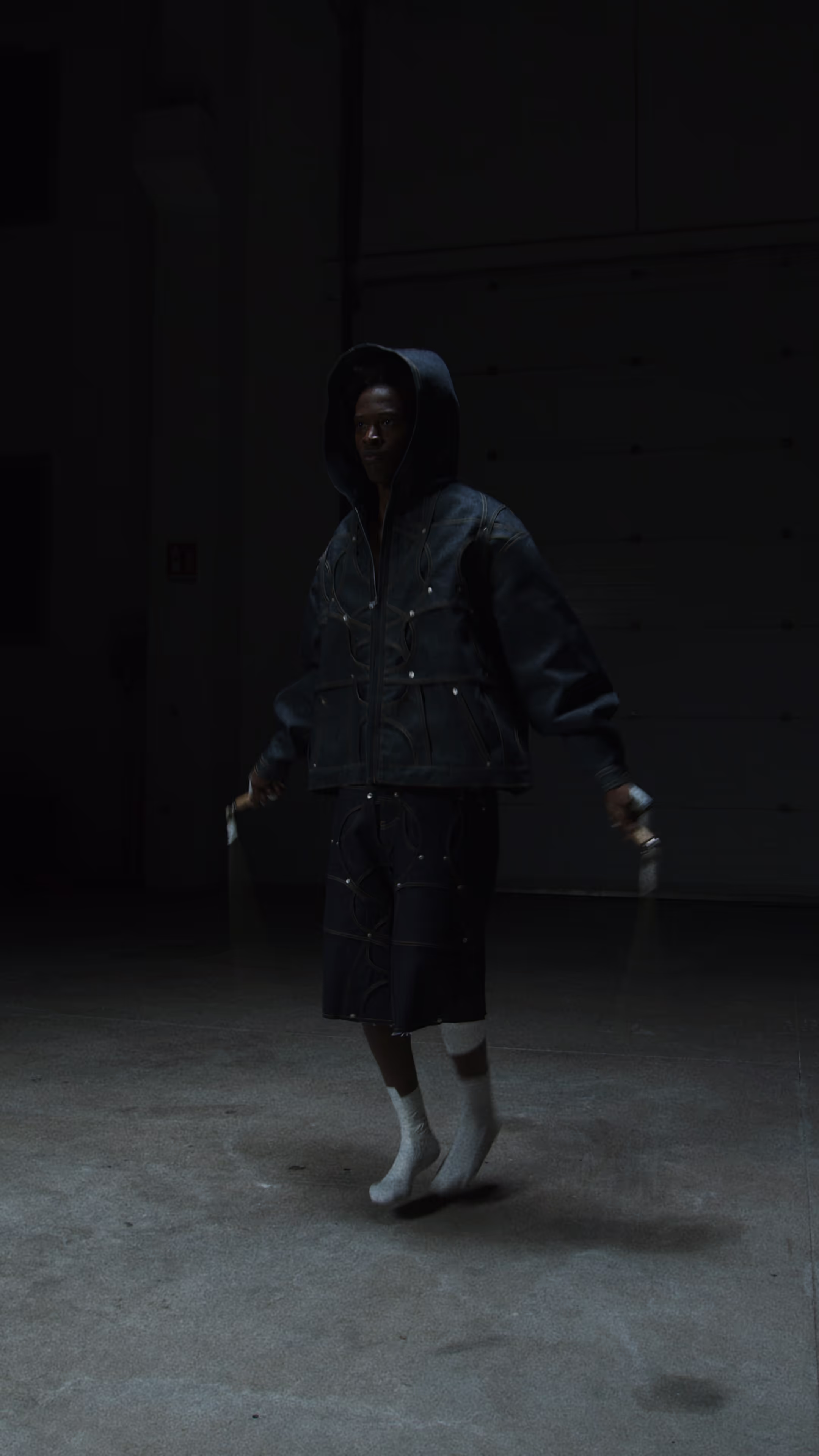 Person wearing a dark oversized hooded jacket and shorts, jumping rope in a dimly lit industrial space.
