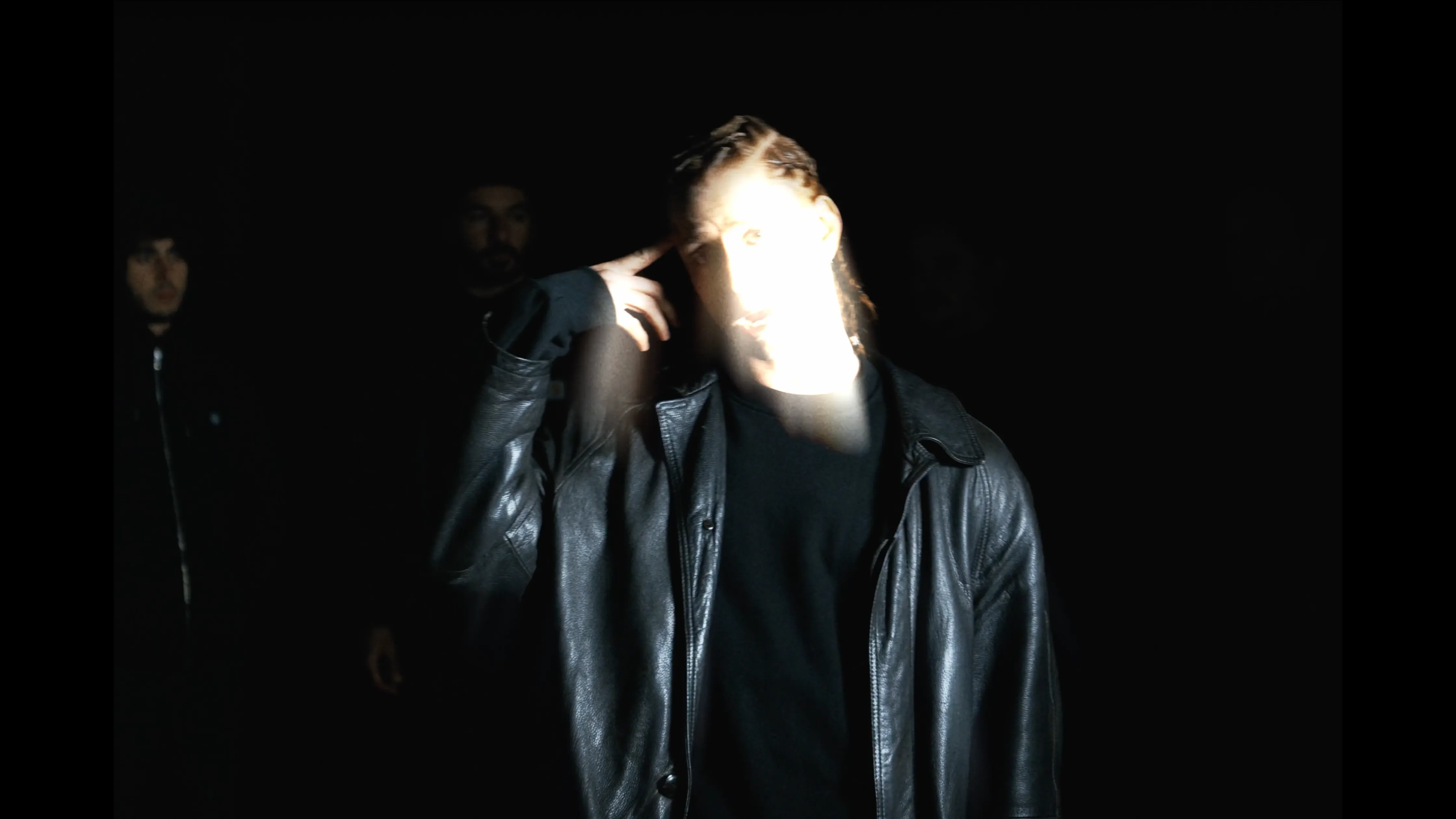 Anelz in black leather jacket pointing to his temple with his right hand, illuminated against a dark background with faint figures behind.
