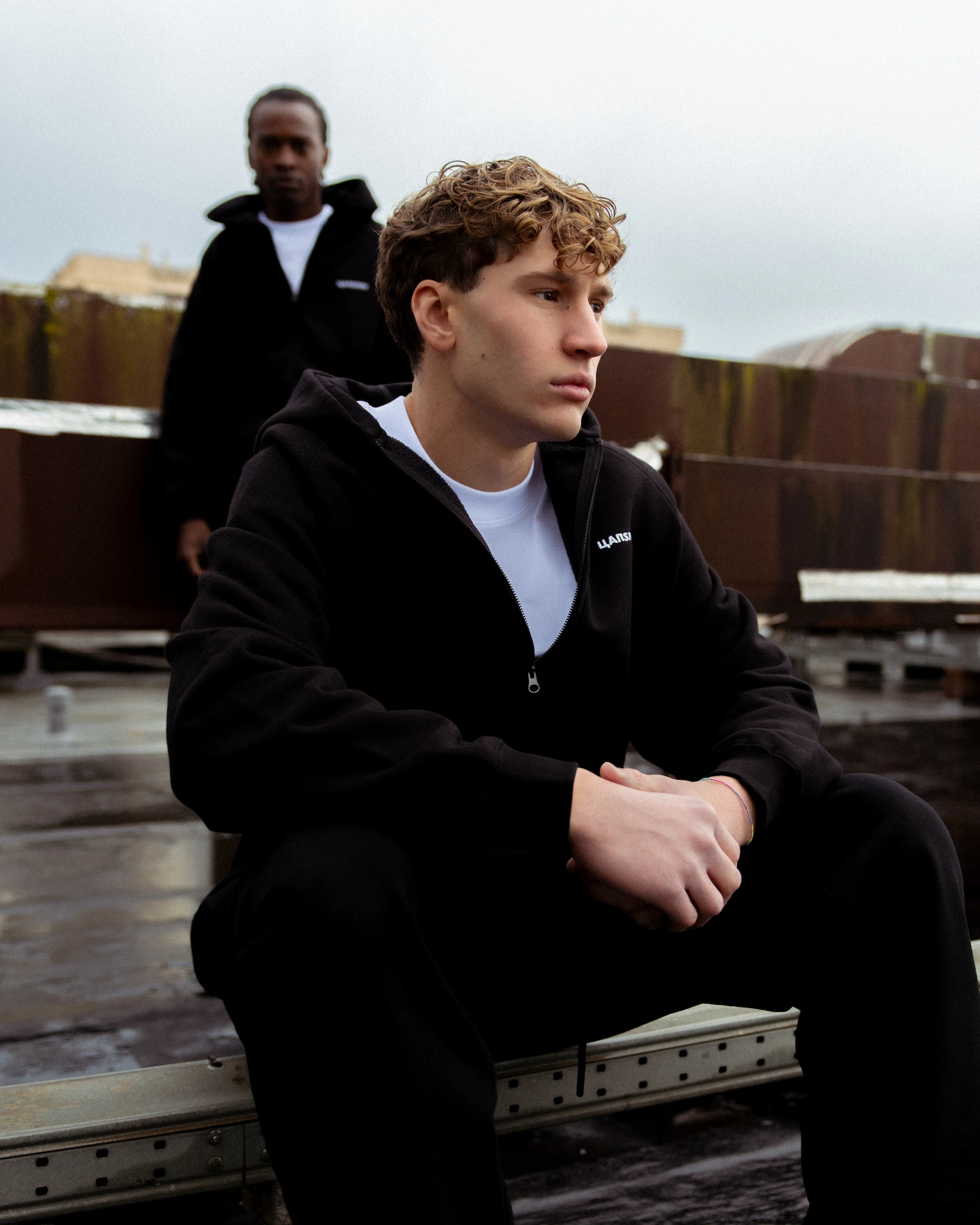 Young man with curly hair in black hoodie sitting pensively on a rooftop with another person blurred in the background.