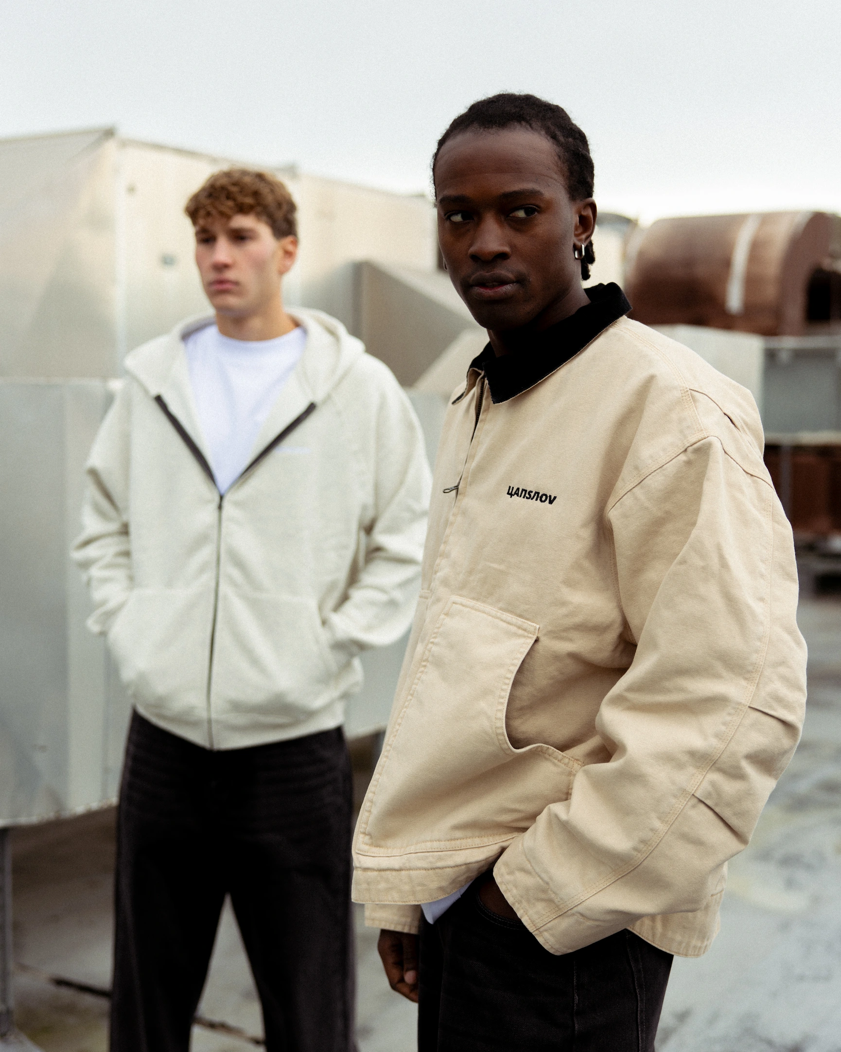 Two young men standing outdoors near industrial equipment wearing casual jackets, one in beige and one in off-white.