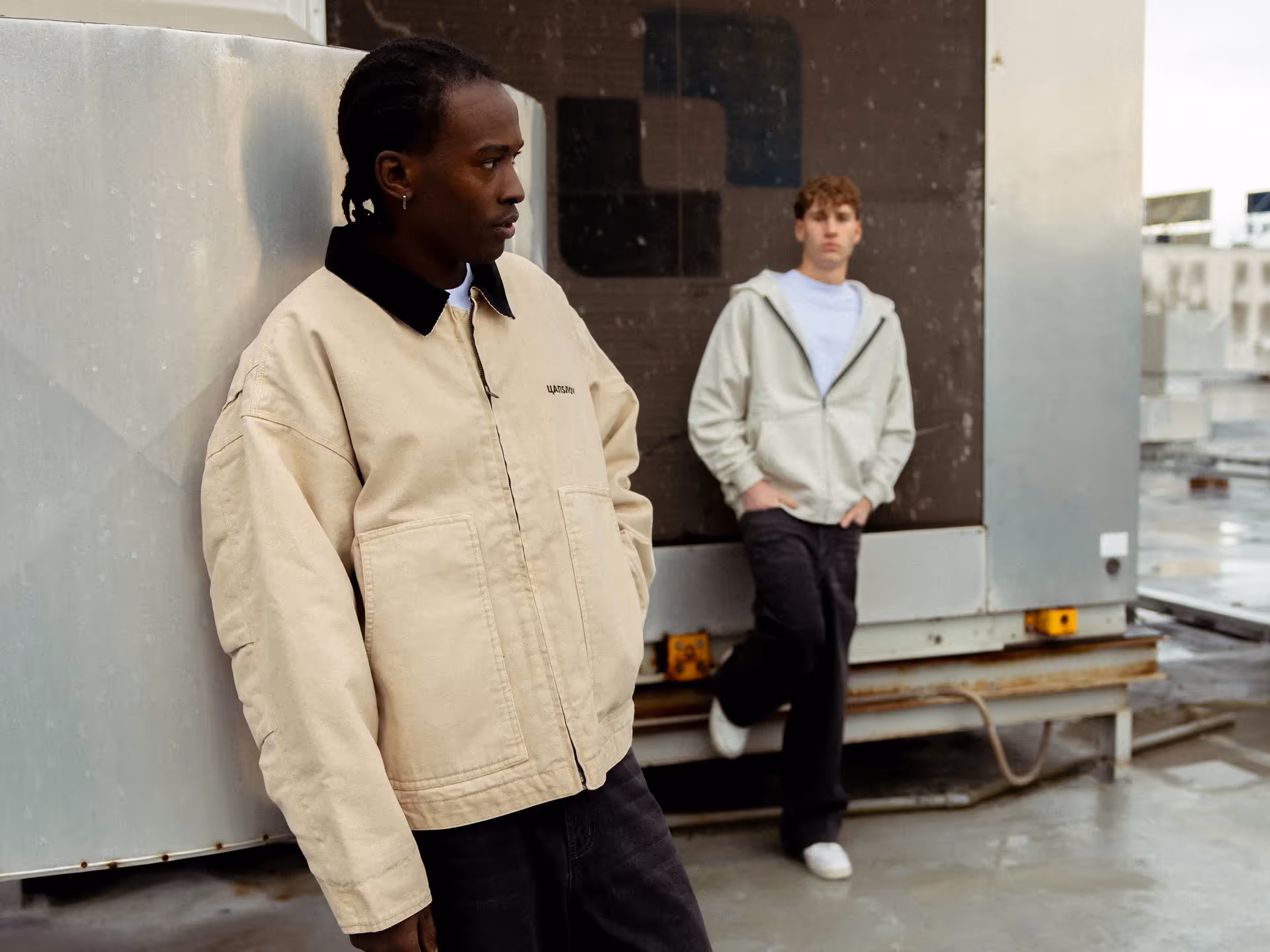 Two young men wearing casual jackets standing on an industrial rooftop with metal structures.