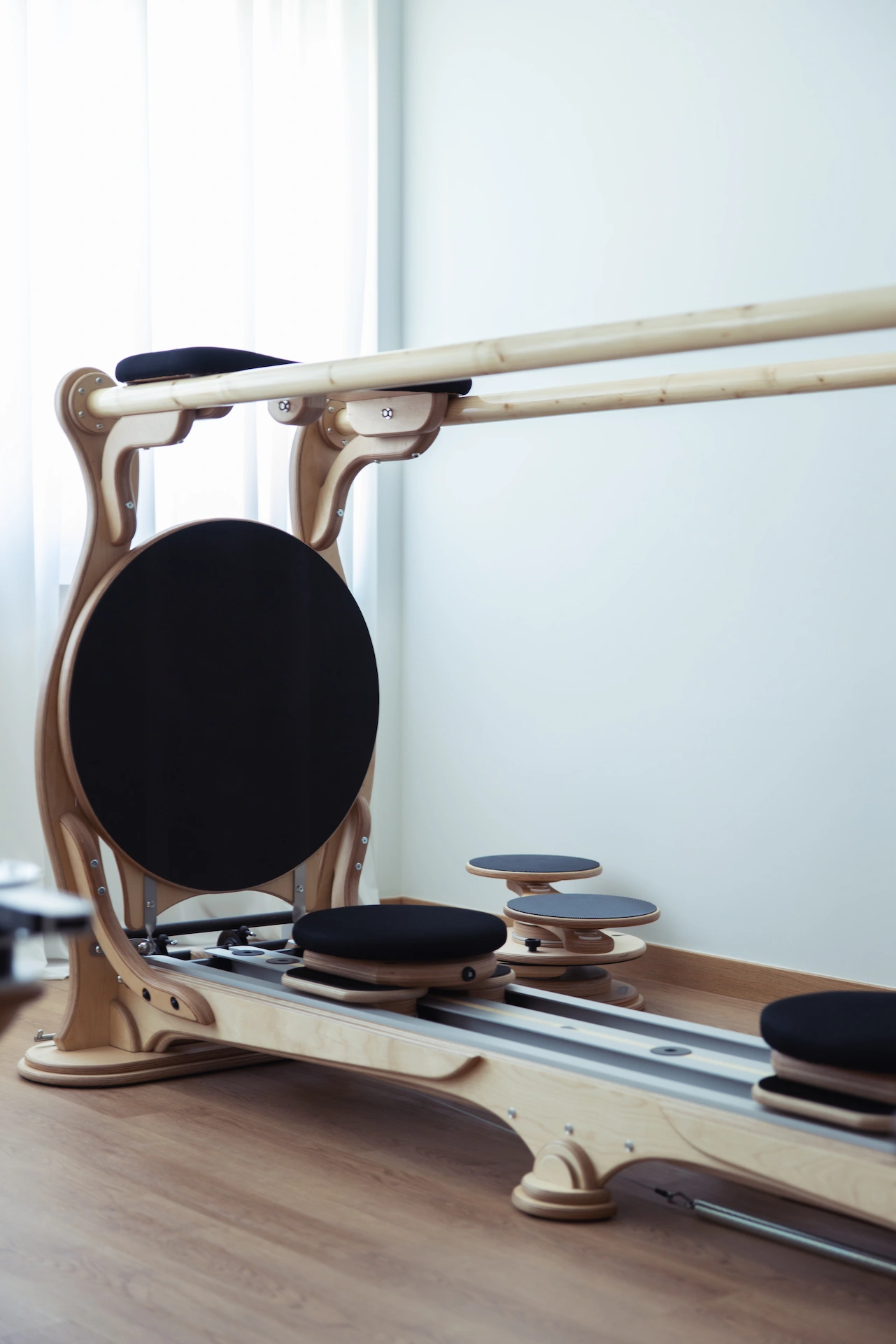 Wooden Pilates reformer machine with black cushions in a bright room with wooden floor.