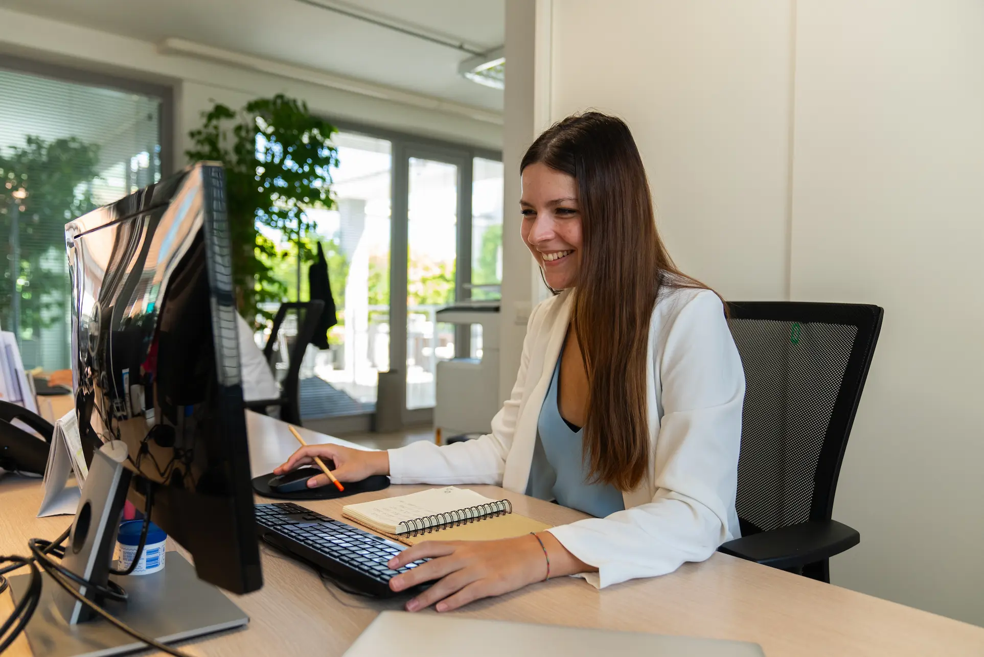 Woman in white blazer smiling and working on a desktop computer in a bright office.