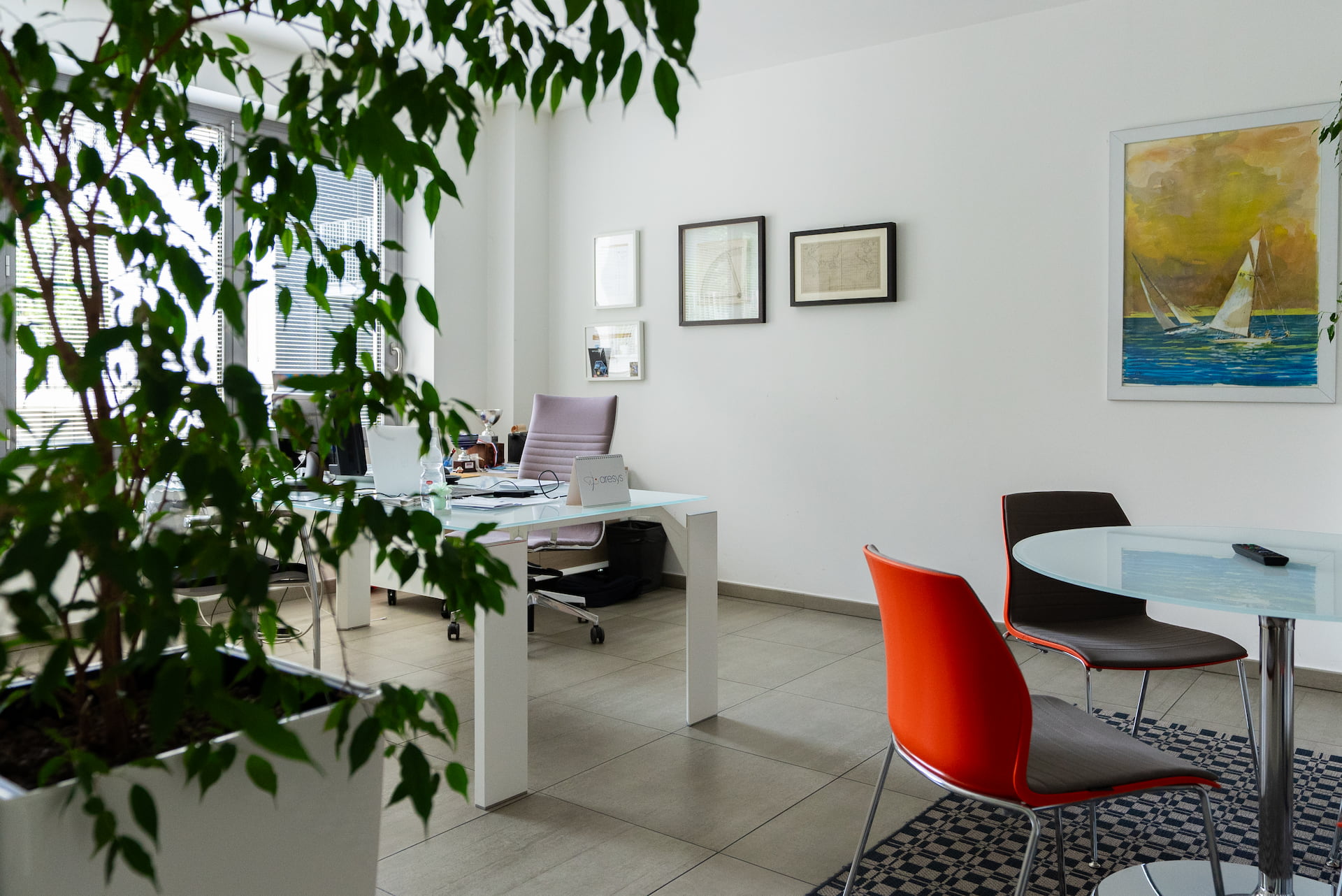 Modern office space with a glass desk, ergonomic chair, potted plant, and a round table with orange and brown chairs on a patterned rug.