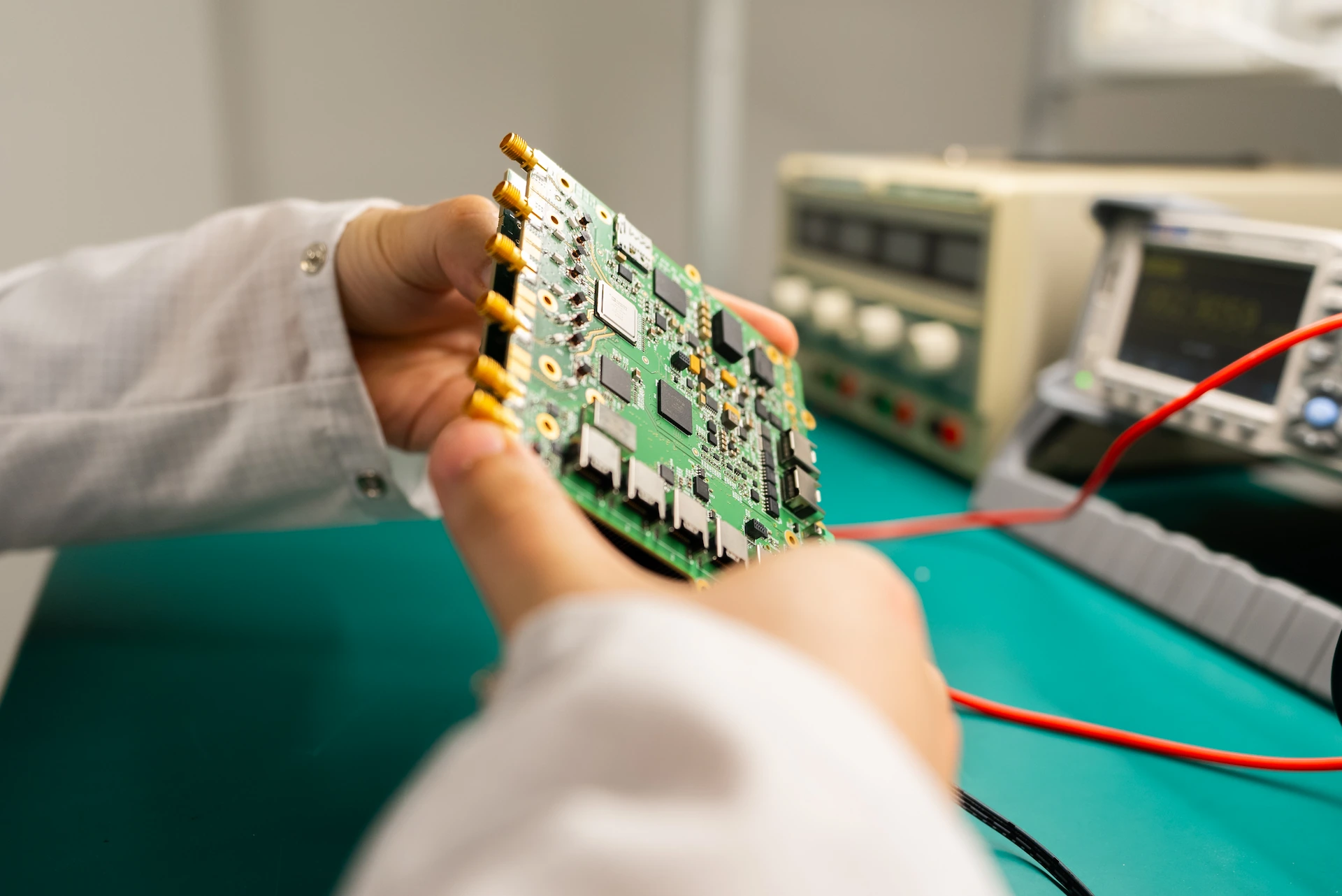 Hands holding a green printed circuit board connected to red test cables in an electronics lab.