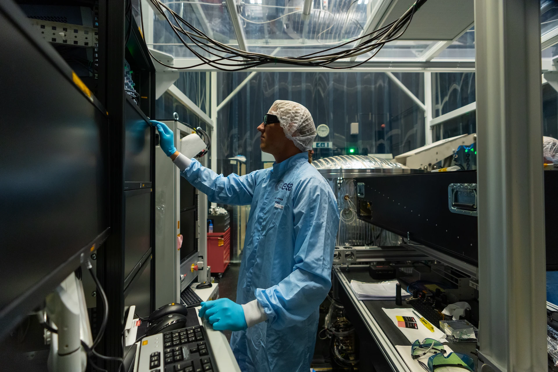 Scientist in a cleanroom suit and hairnet operating scientific equipment with multiple monitors and wires in a laboratory.
