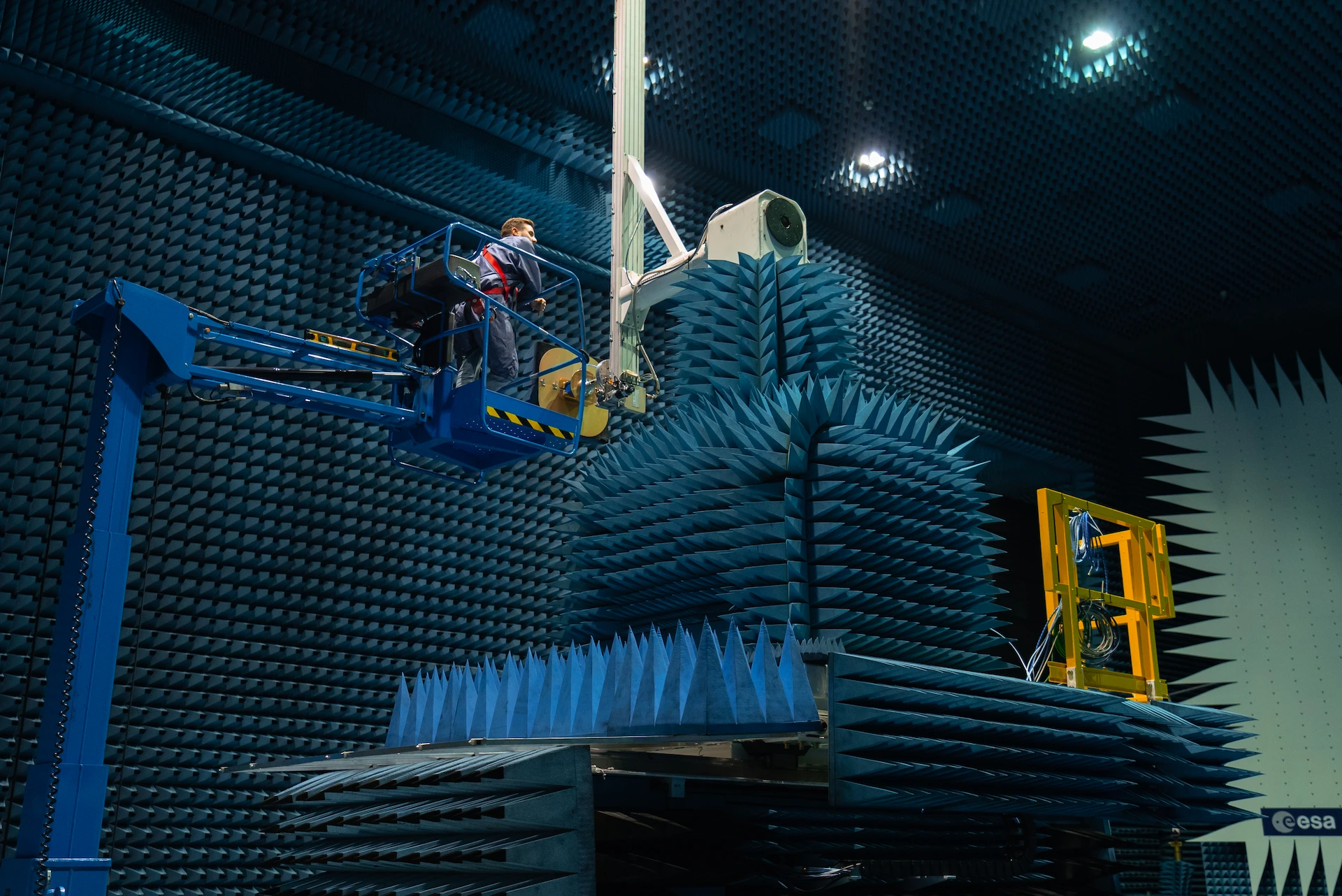 Technician on a blue lift inside an anechoic chamber testing a large antenna covered with blue foam pyramids.