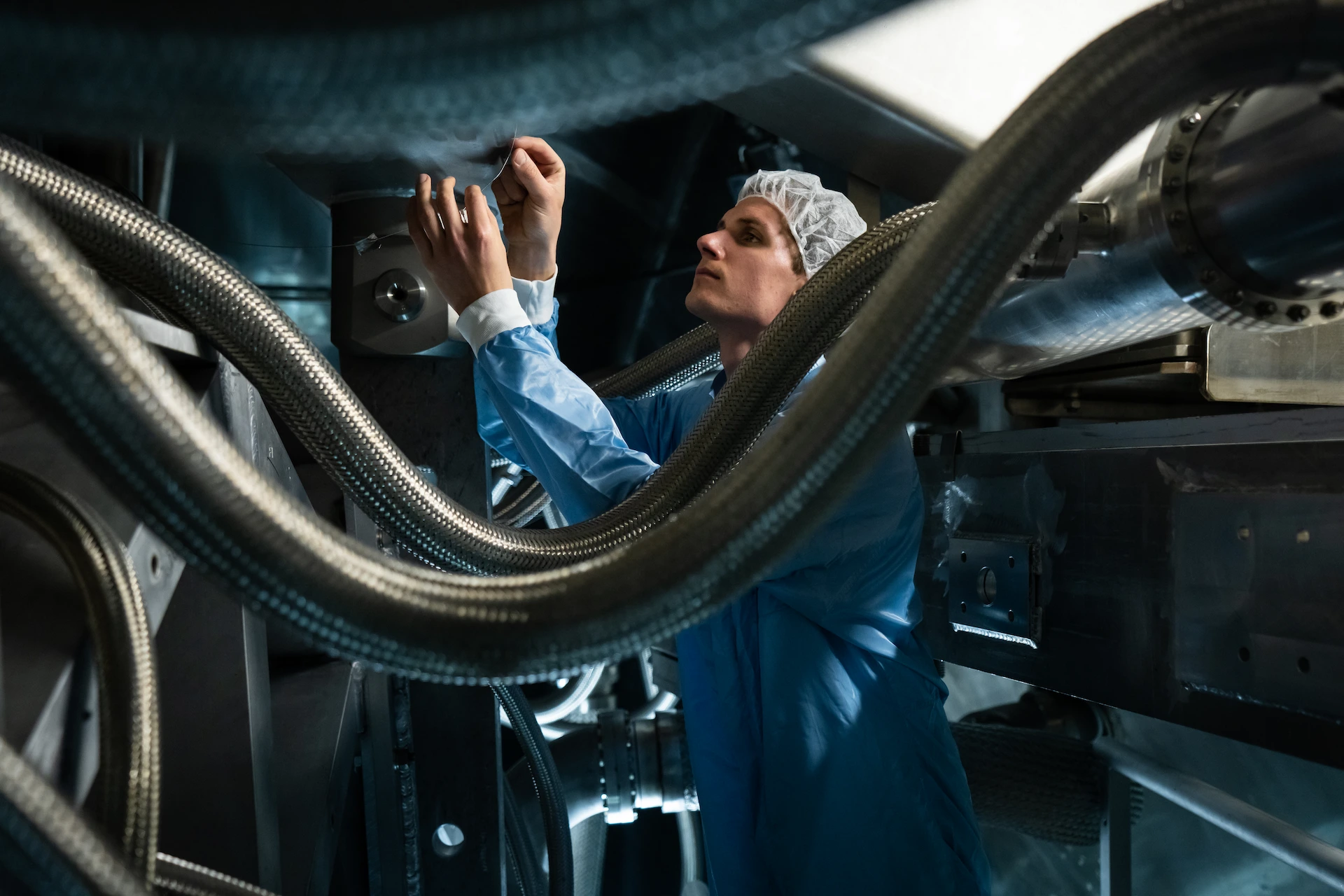 Human in cleanroom suit inspecting or assembling metal machinery with flexible hoses.
