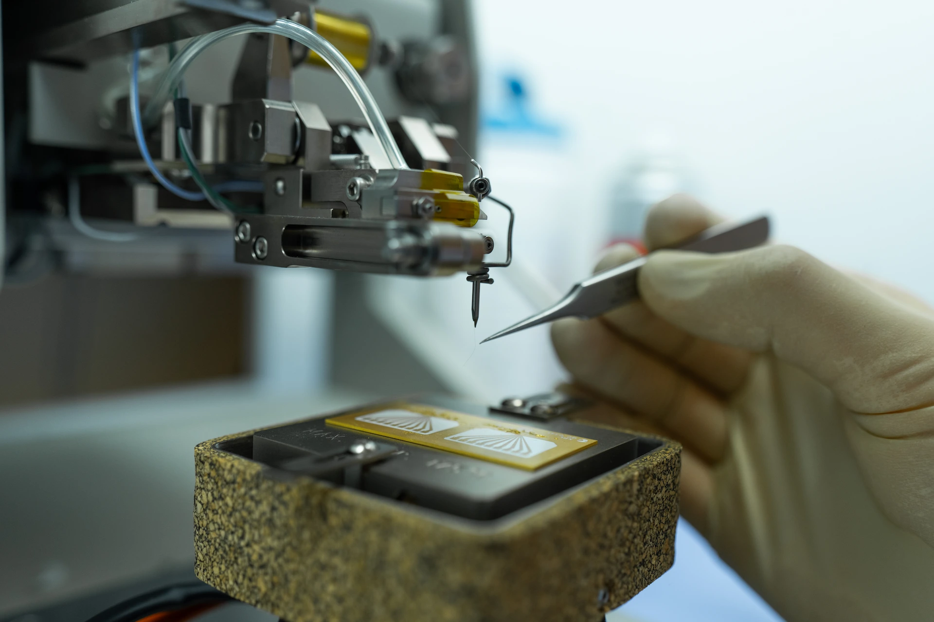 Gloved hand using tweezers to manipulate a delicate component on a precision machine.