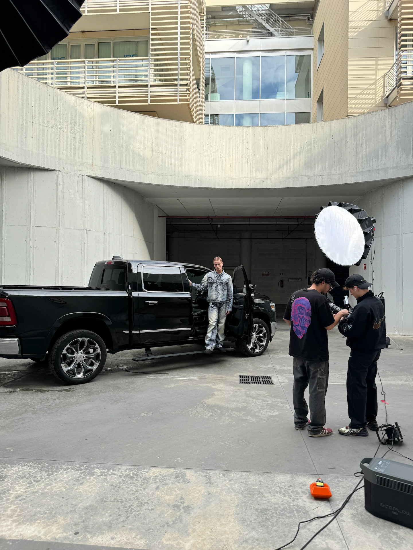 Model in denim outfit standing in the open door of a black pickup truck during a photoshoot with two crew members checking equipment nearby.