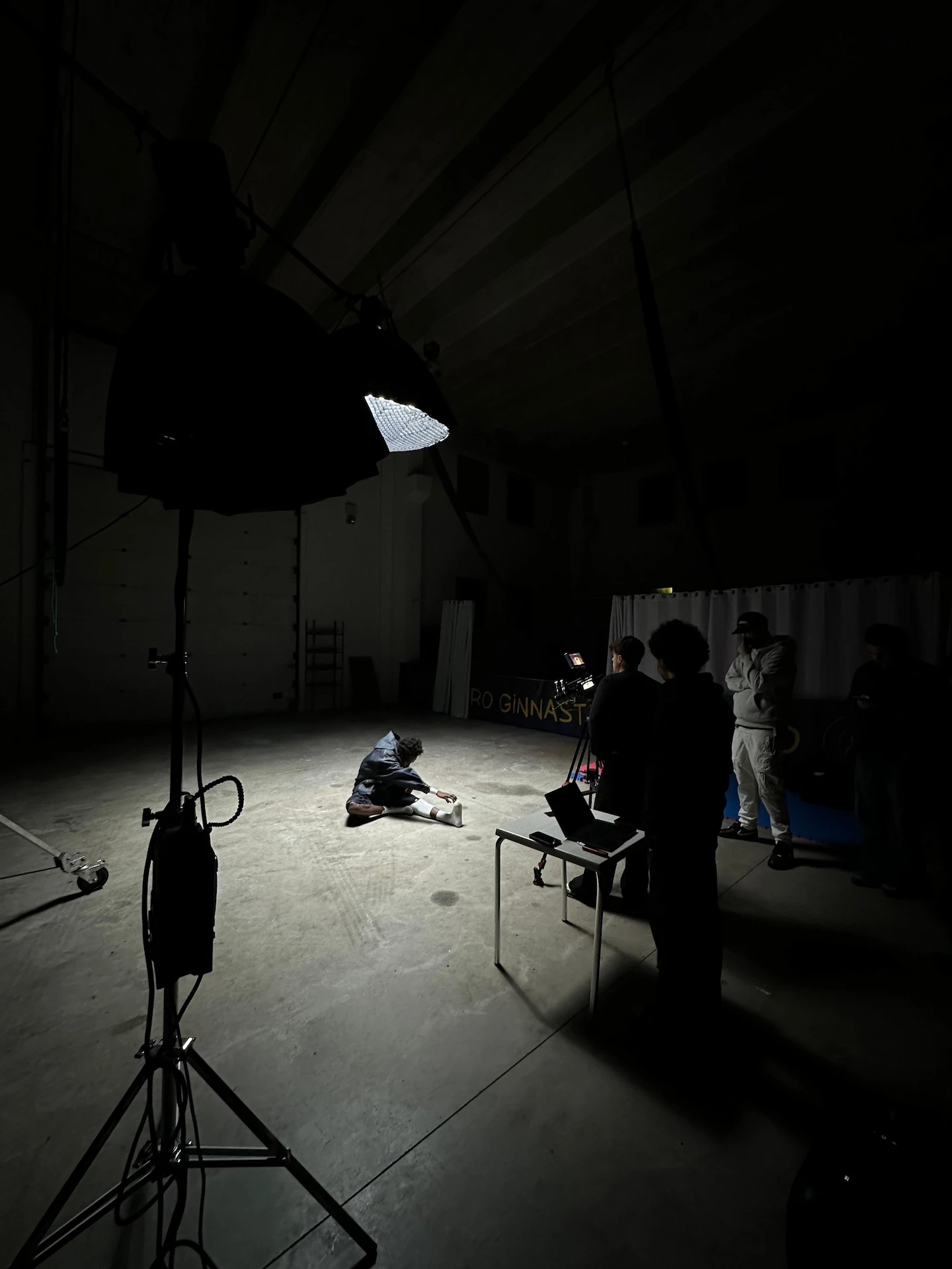Person stretching on floor under a large studio light with a small film crew operating camera and laptop in a dark warehouse.