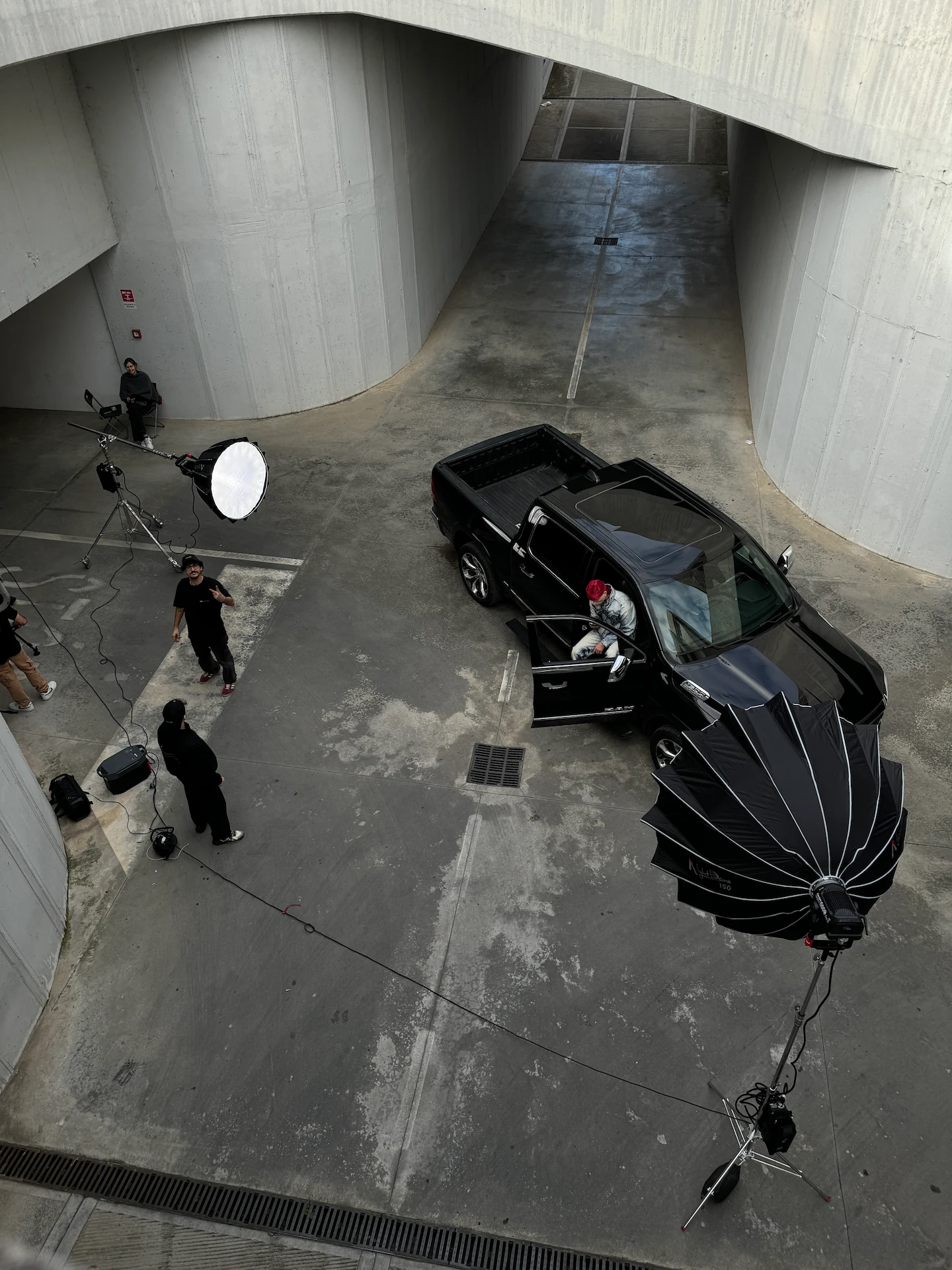Photo shoot setup with a black pickup truck parked in an urban concrete area, surrounded by large photography lights and several people.