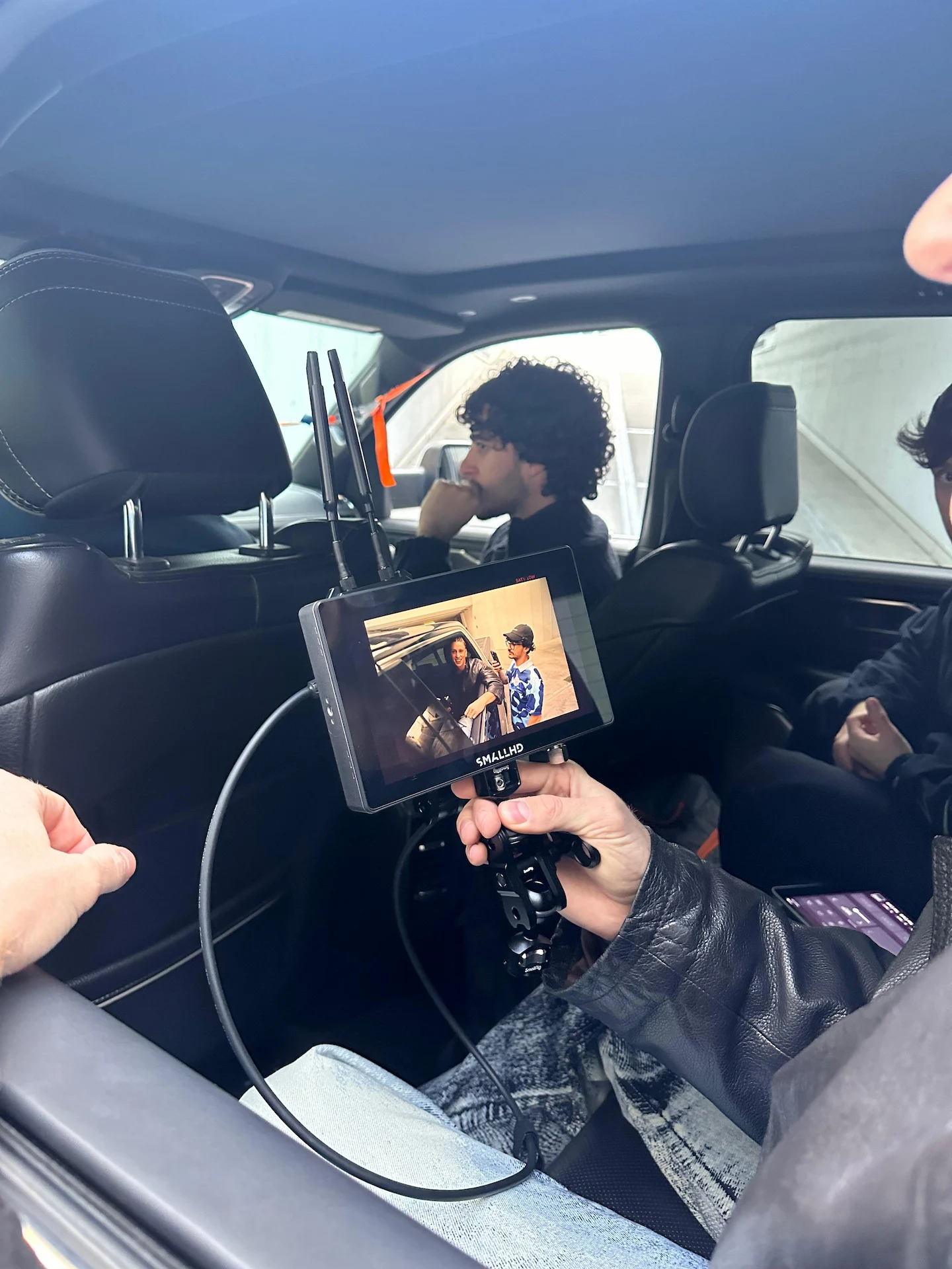 Person inside a car holding a SmallHD monitor displaying two people talking by a vehicle, with another person sitting in the back seat.