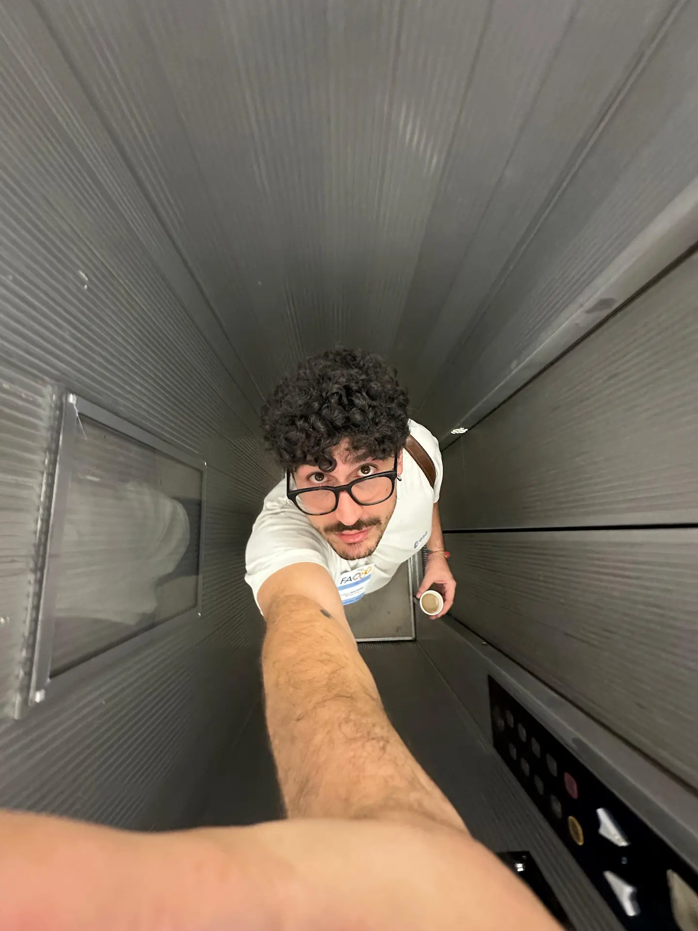 Young man with curly hair and glasses taking a selfie in a narrow elevator holding a cup.