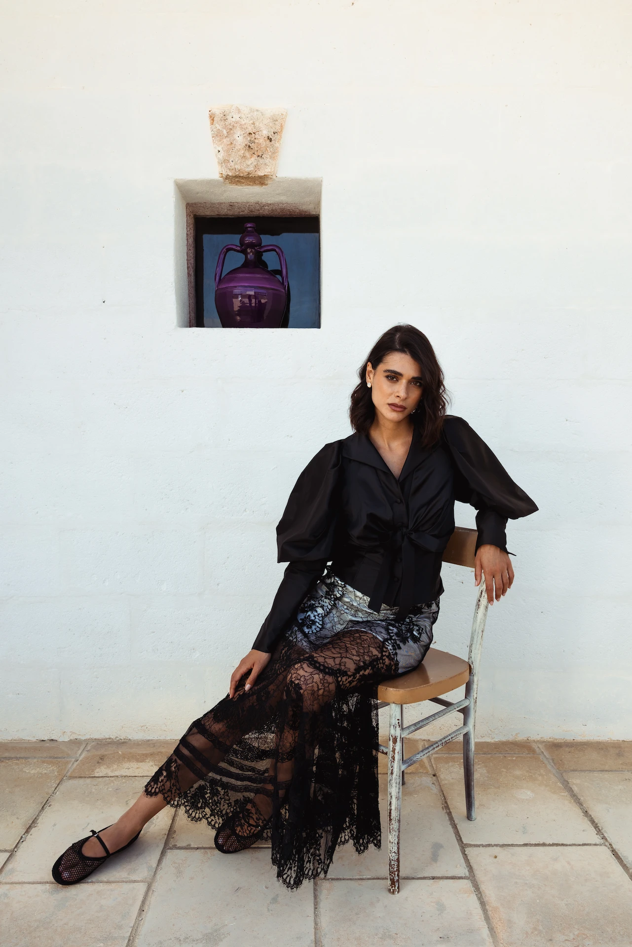 Woman sitting on a wooden chair in front of a white wall, wearing a black blouse and a black sheer lace skirt.