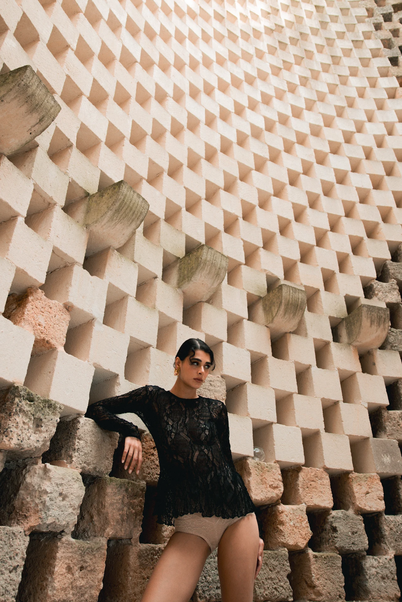 Woman wearing a black lace top and beige bottoms standing against a textured wall made of protruding white and rustic stone blocks.