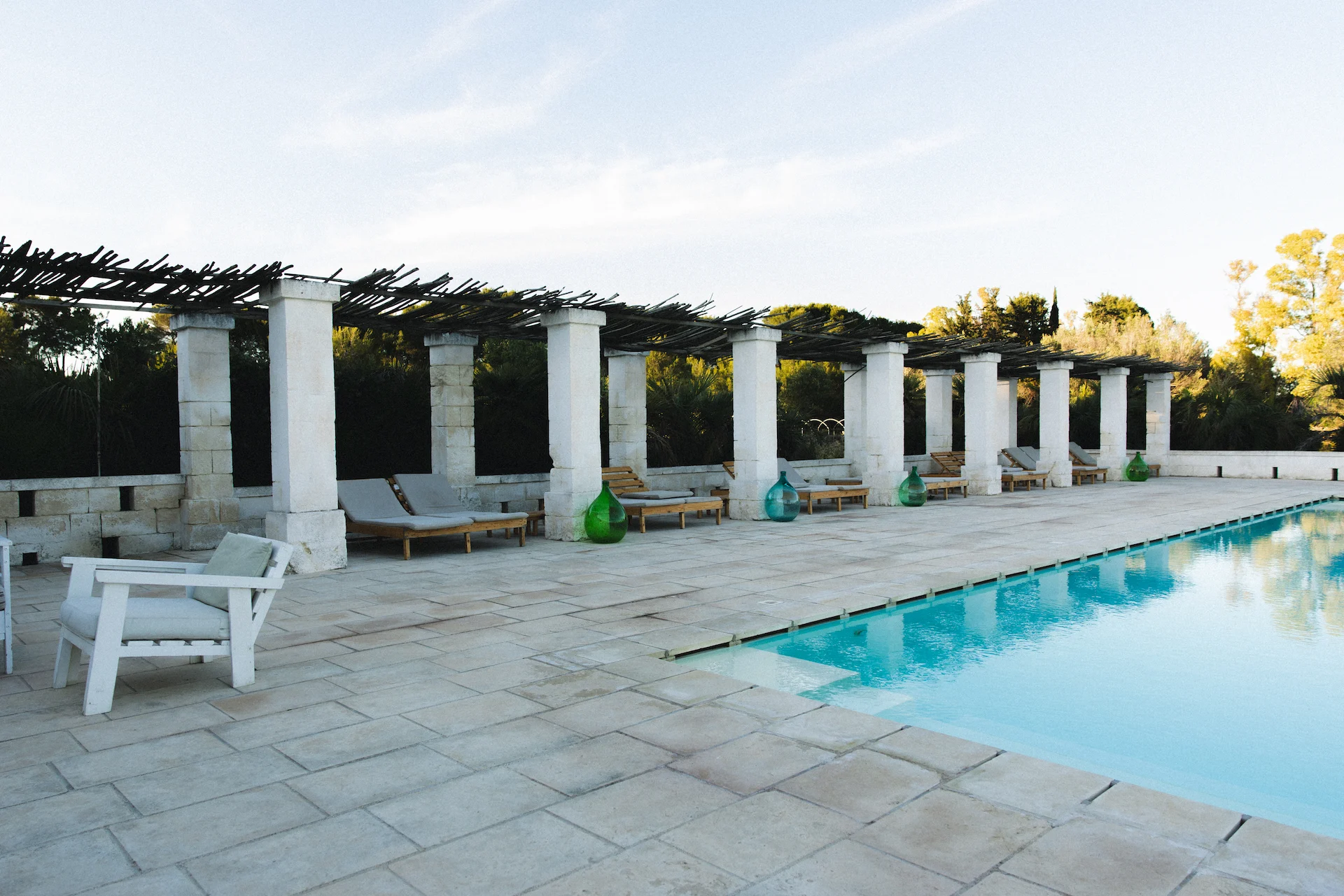 Outdoor swimming pool with stone tile deck, lounge chairs under wooden pergolas, and green and blue glass decorative vases.