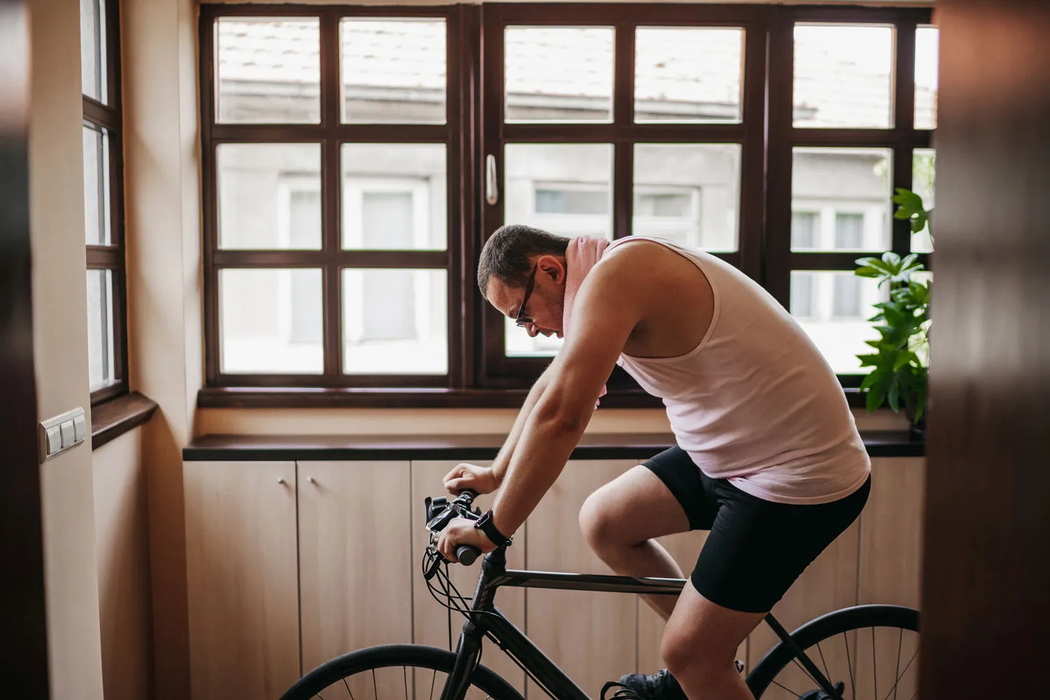 Man wearing a tank top and cycling shorts riding a bicycle indoors in front of a window.