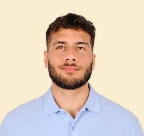 Portrait of a young man with short curly hair and a beard, wearing a light blue polo shirt, against a beige background.