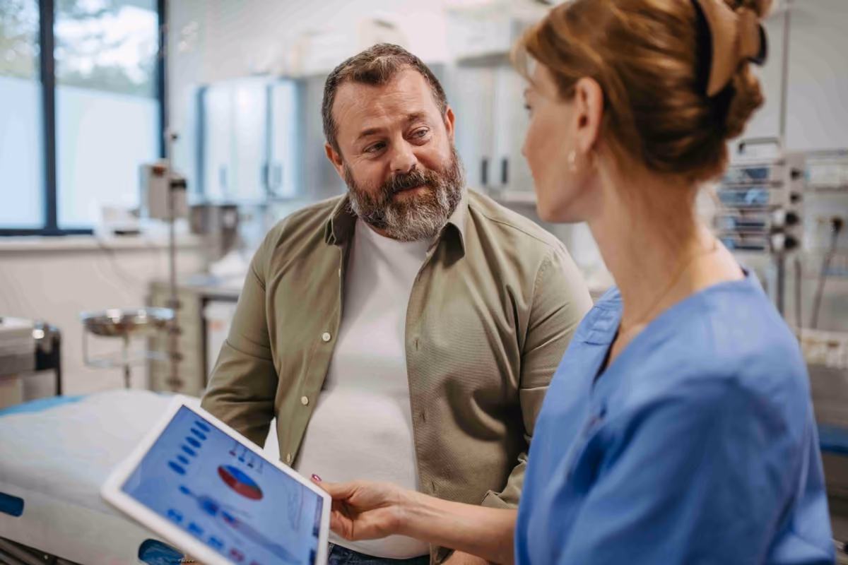 Male patient with beard in clinic looking at female nurse showing tablet with health charts.