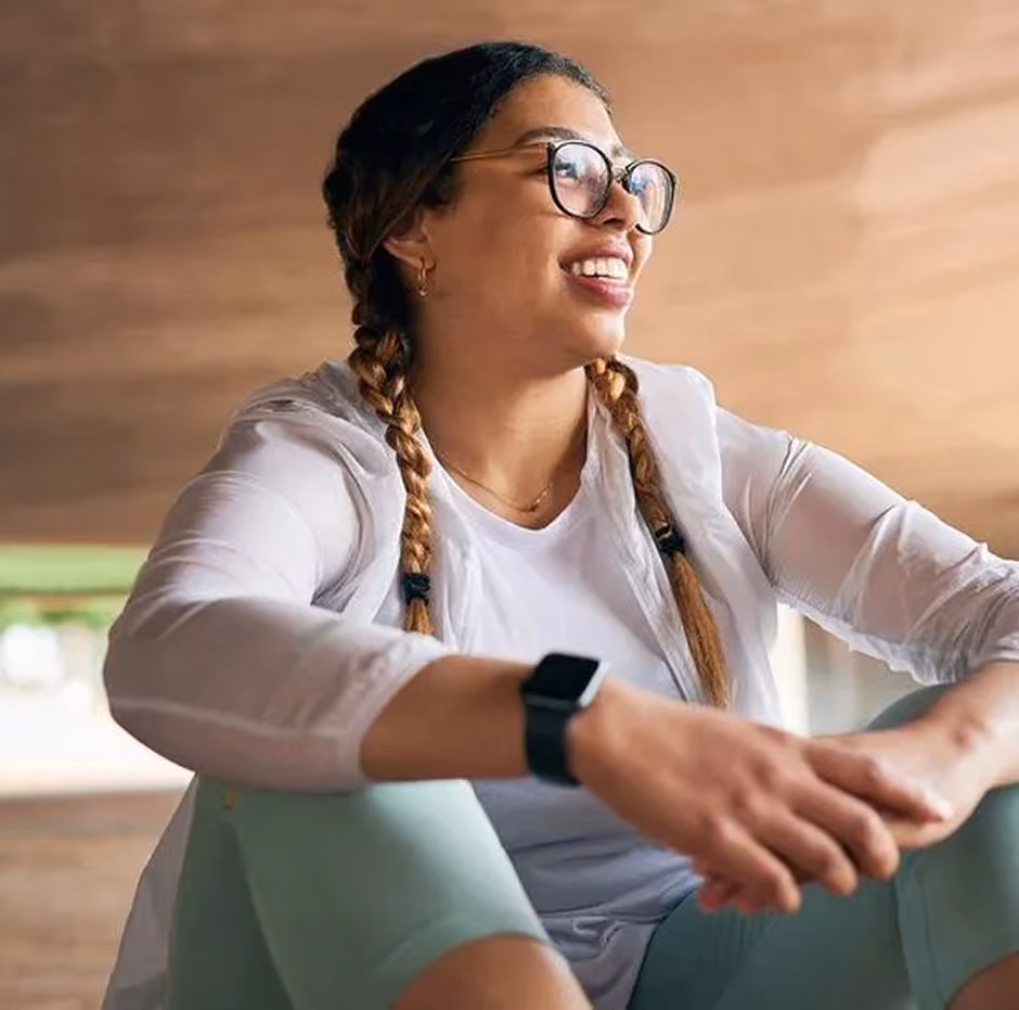 Smiling woman with glasses and braids sits wearing workout clothes and a smartwatch indoors.