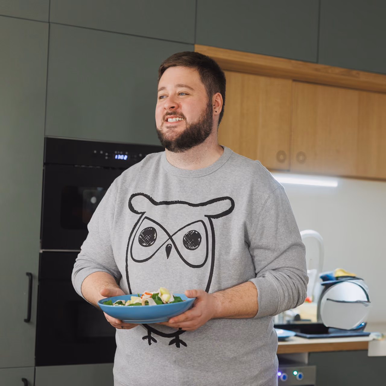 Bearded man smiling in kitchen holding a blue bowl of salad with vegetables.