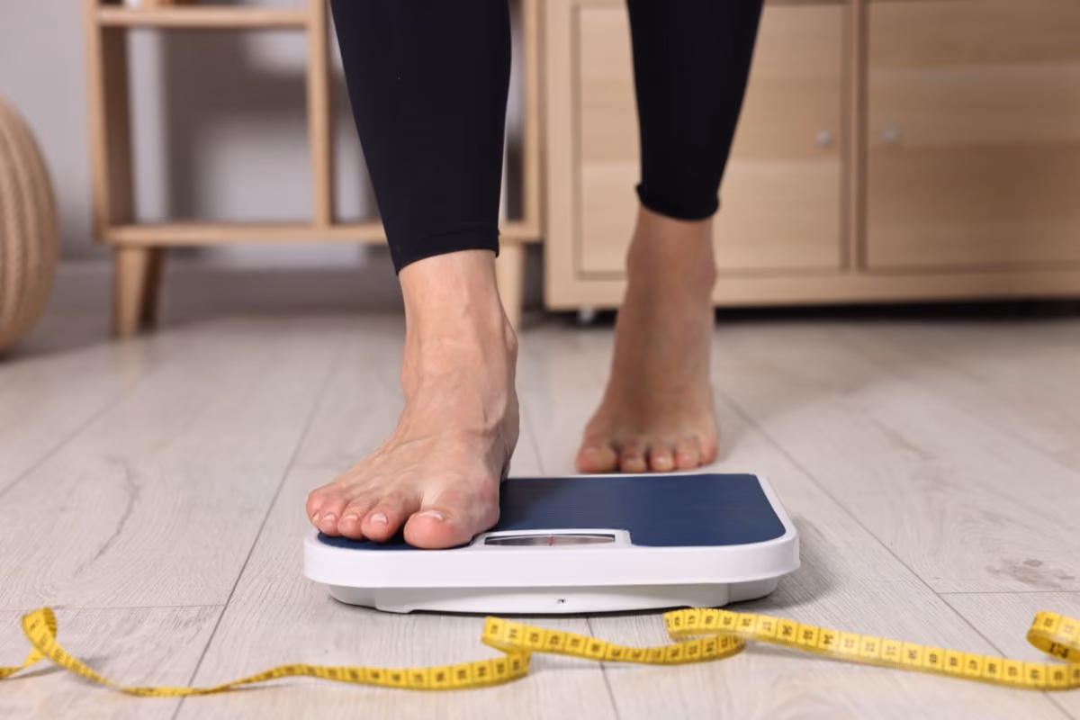 Person stepping onto a bathroom scale on a wooden floor with a yellow measuring tape nearby.