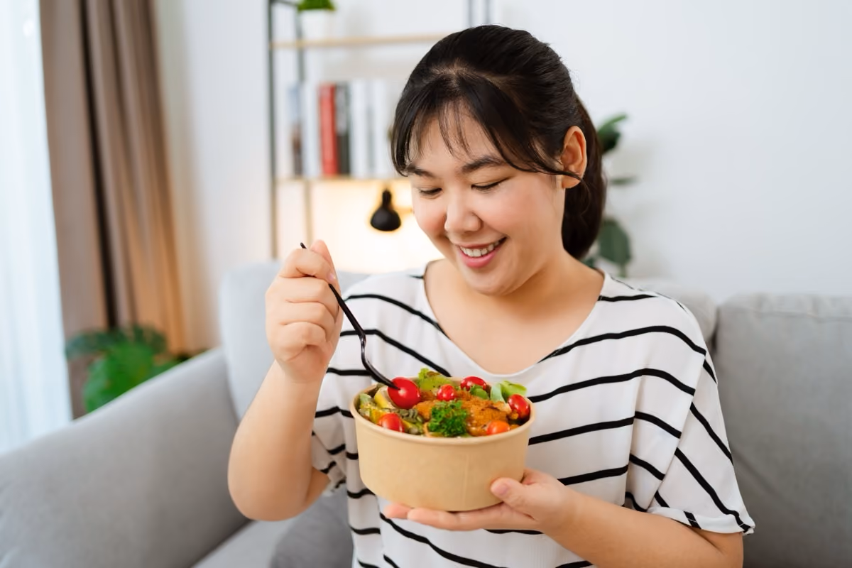 Smiling woman holding a bowl of colorful salad with cherry tomatoes and greens using a black fork indoors.