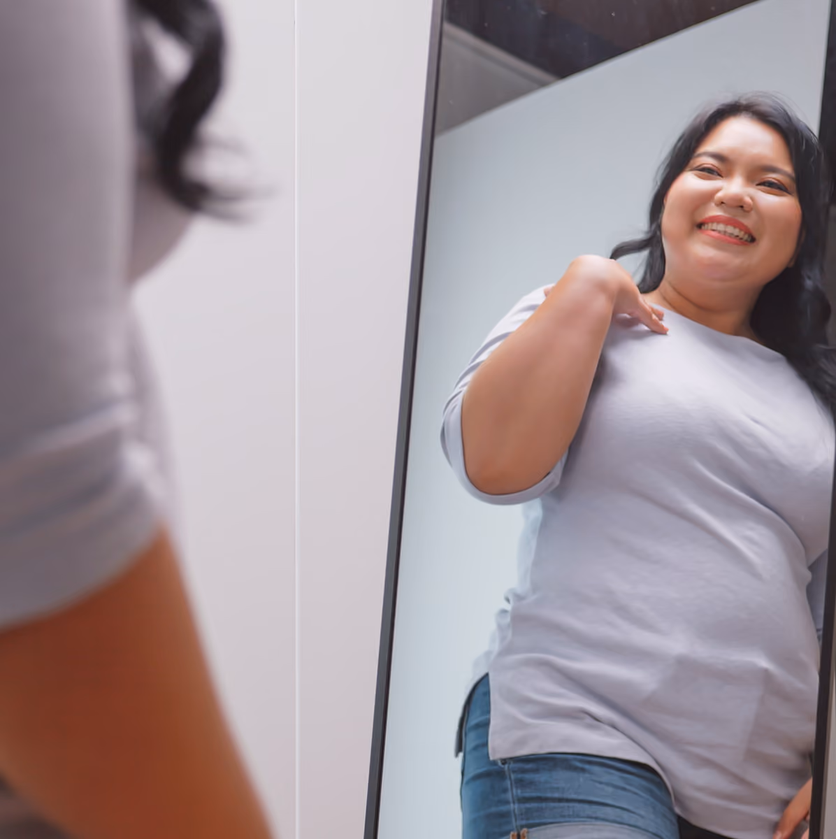 Smiling woman looking at herself in a mirror, wearing a gray shirt and jeans.
